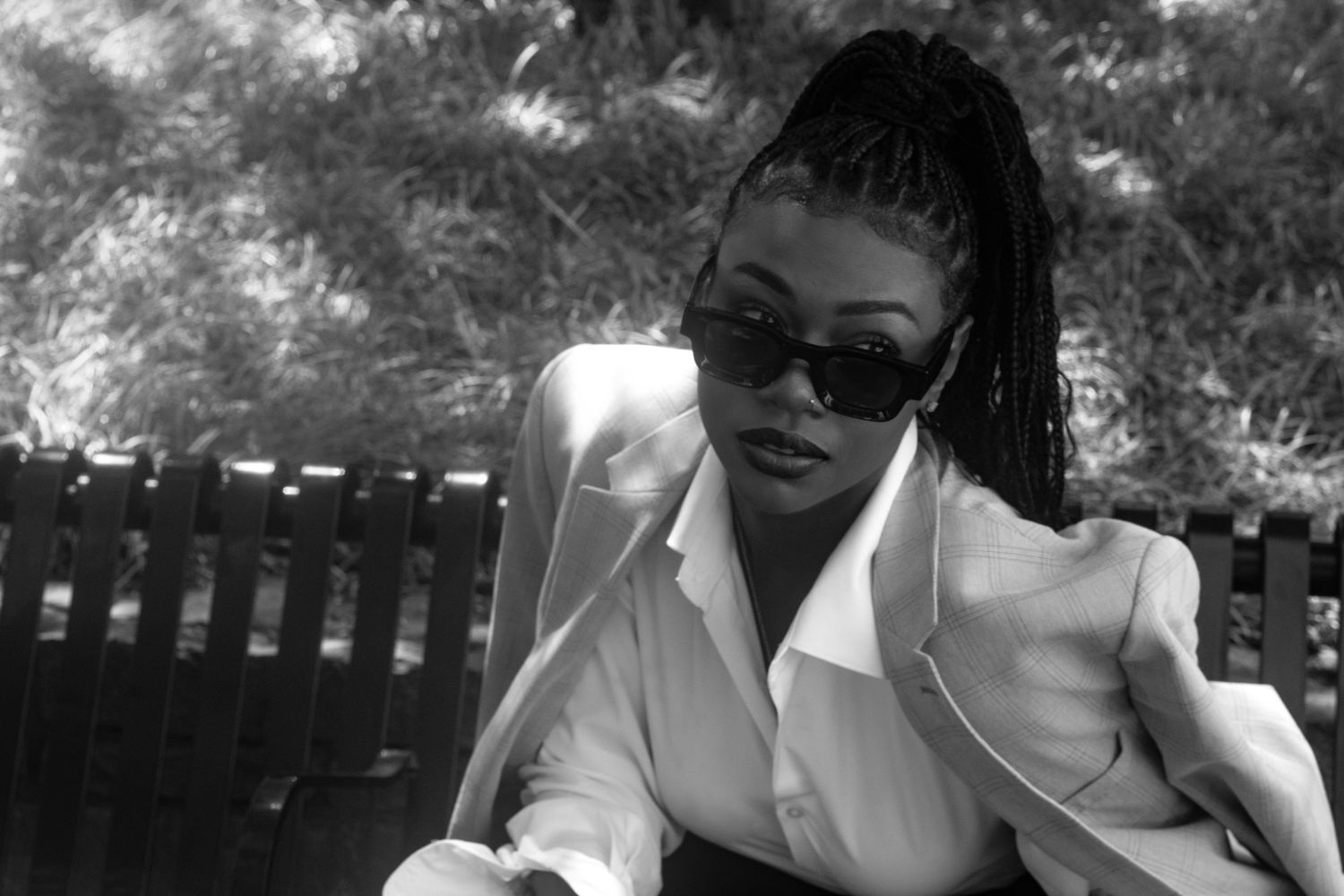Black and white photo of a stylish woman with braids wearing sunglasses and a white blazer seated outdoors.