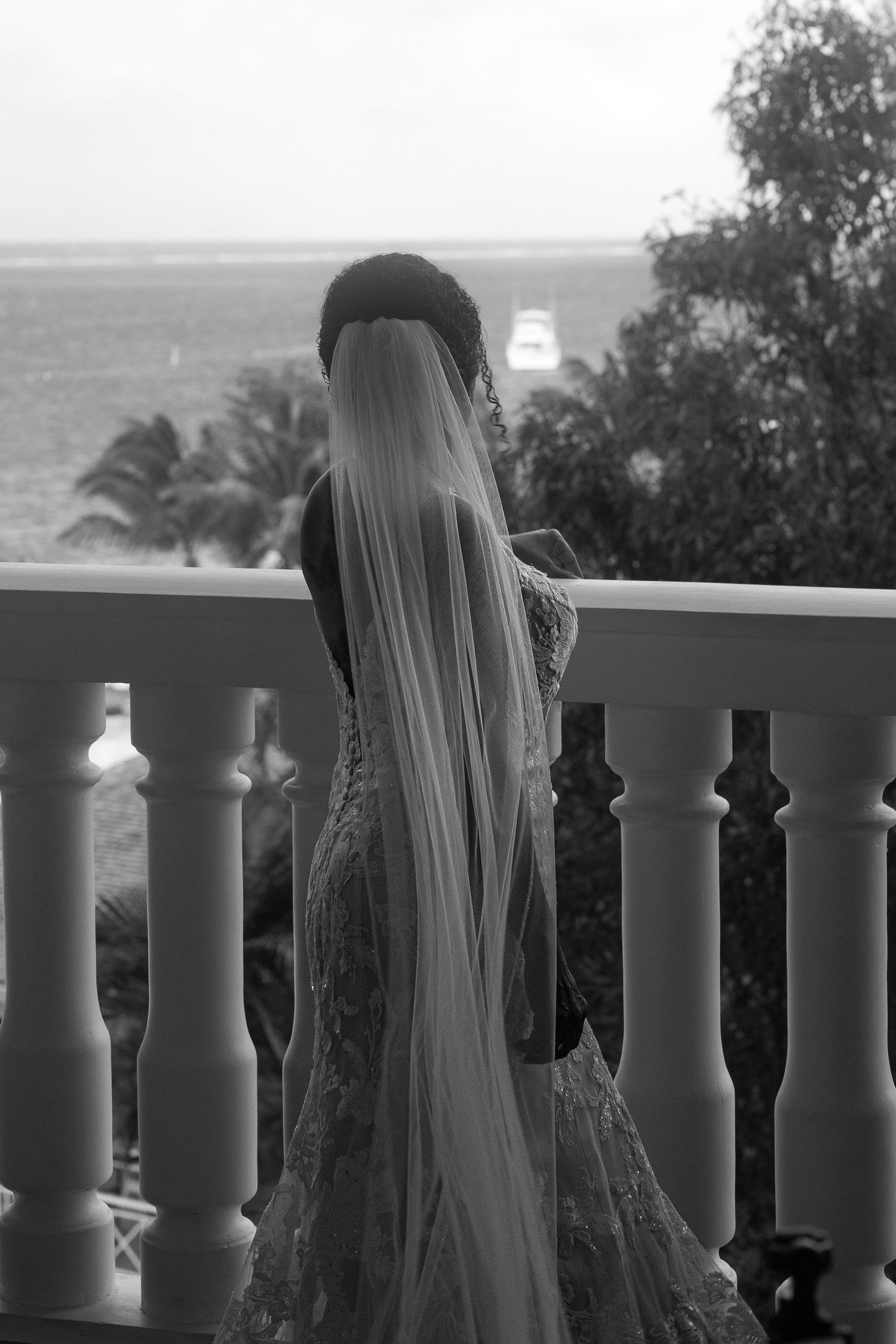 Black and white photo of bride in lace gown and long veil standing on balcony overlooking ocean and palm trees.