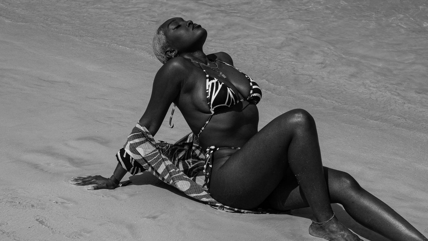 Black and white photo of a woman in a bikini sitting on a sandy beach, posing gracefully in the sun.