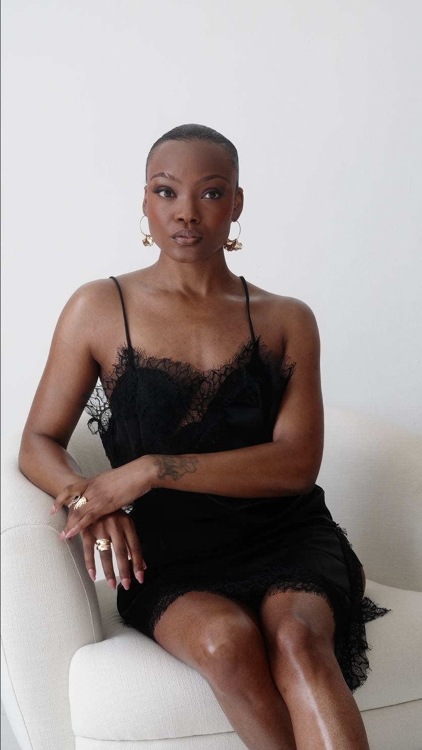 Confident Black woman in black lace dress seated on white chair in minimalist studio setting.
