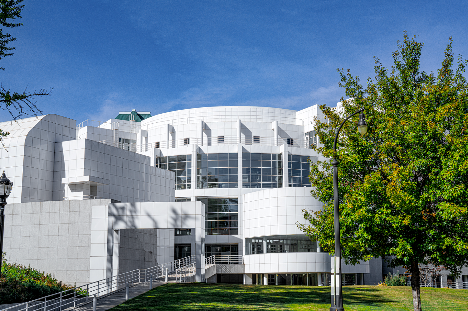 Modern white building with geometric architecture under blue sky surrounded by green trees and manicured lawn.
