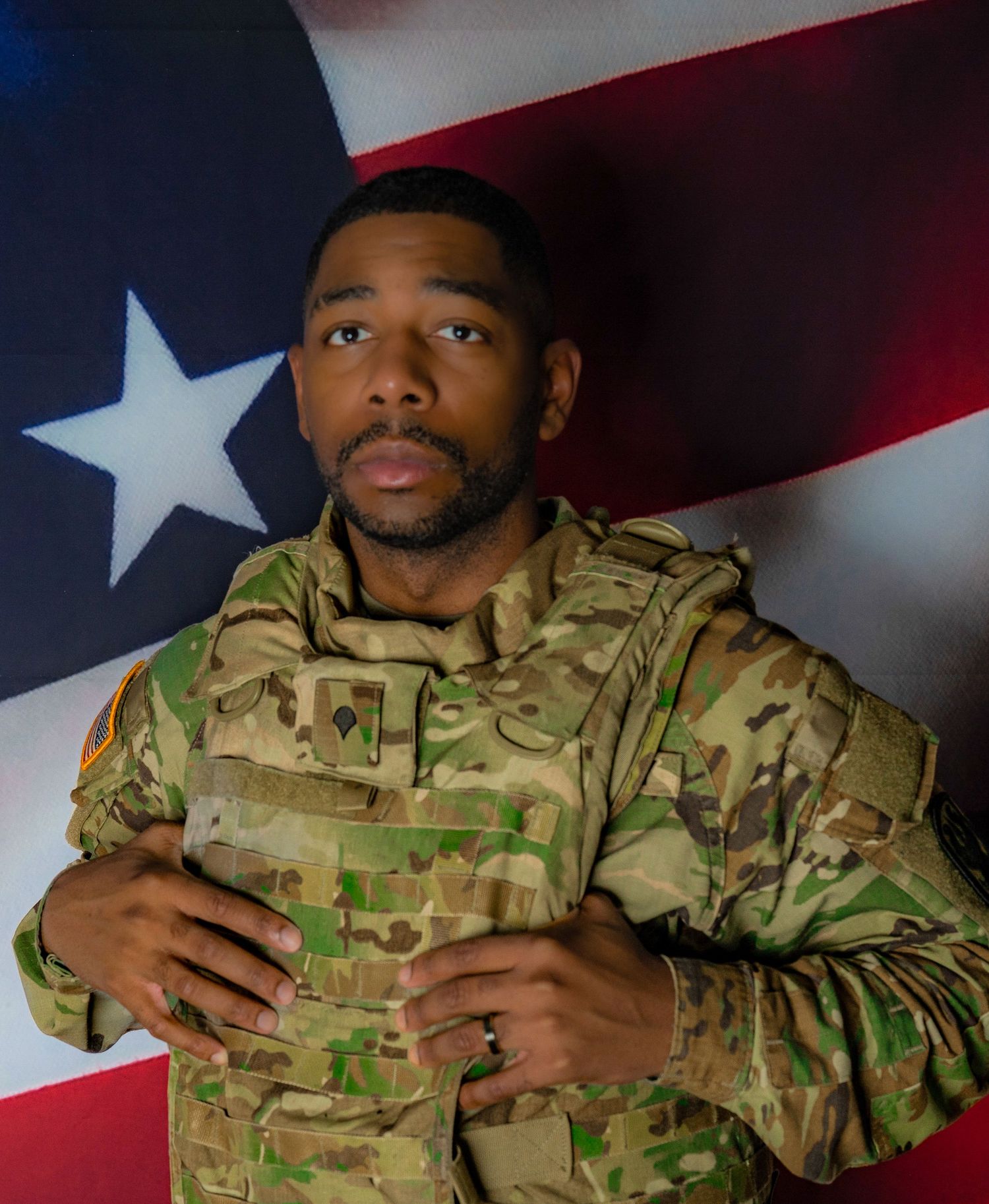 Soldier in military camouflage uniform posing confidently in front of an American flag backdrop.