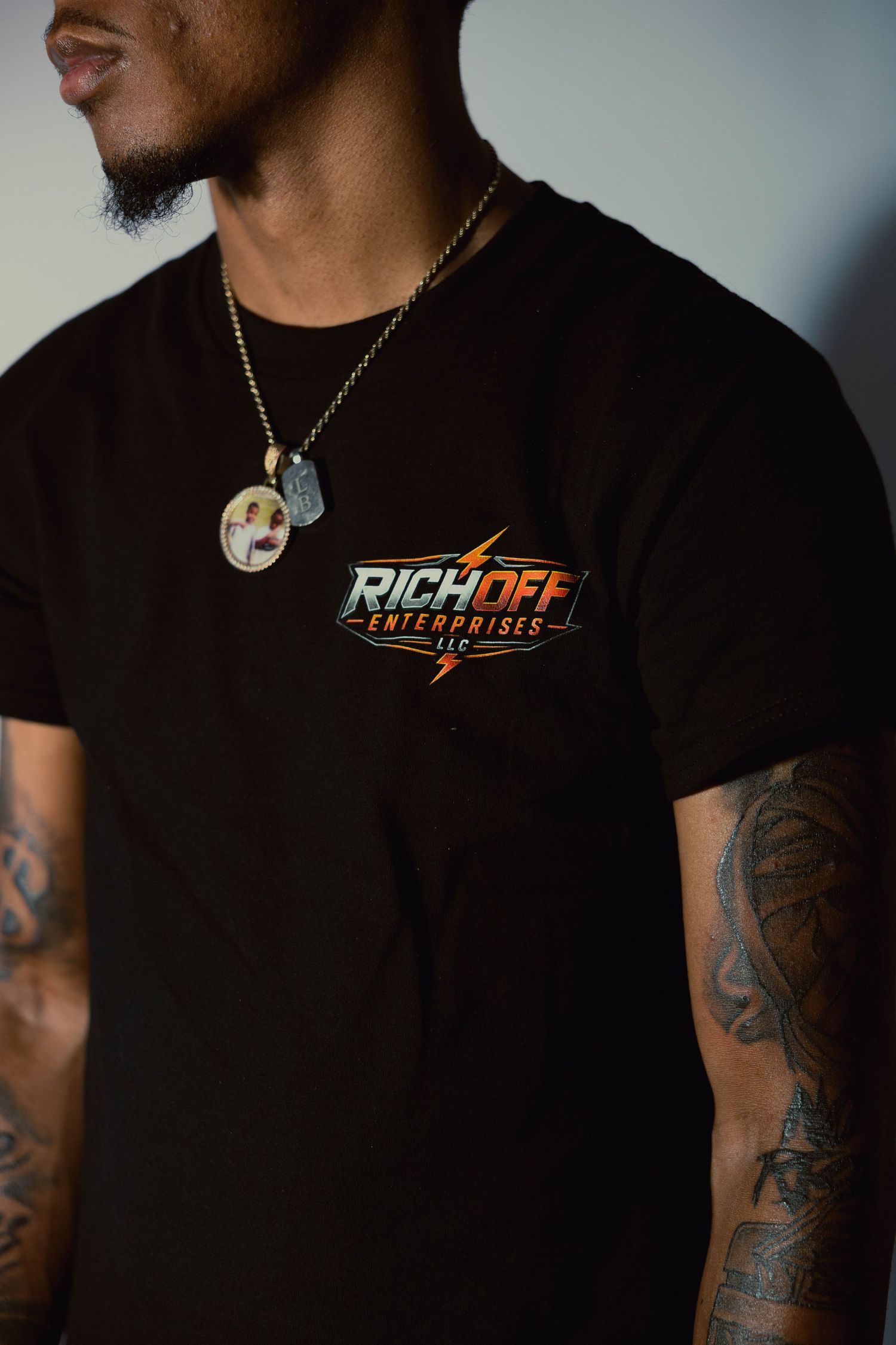 Man wearing a black RichOff Enterprises LLC t-shirt with tattoos visible on his arms.