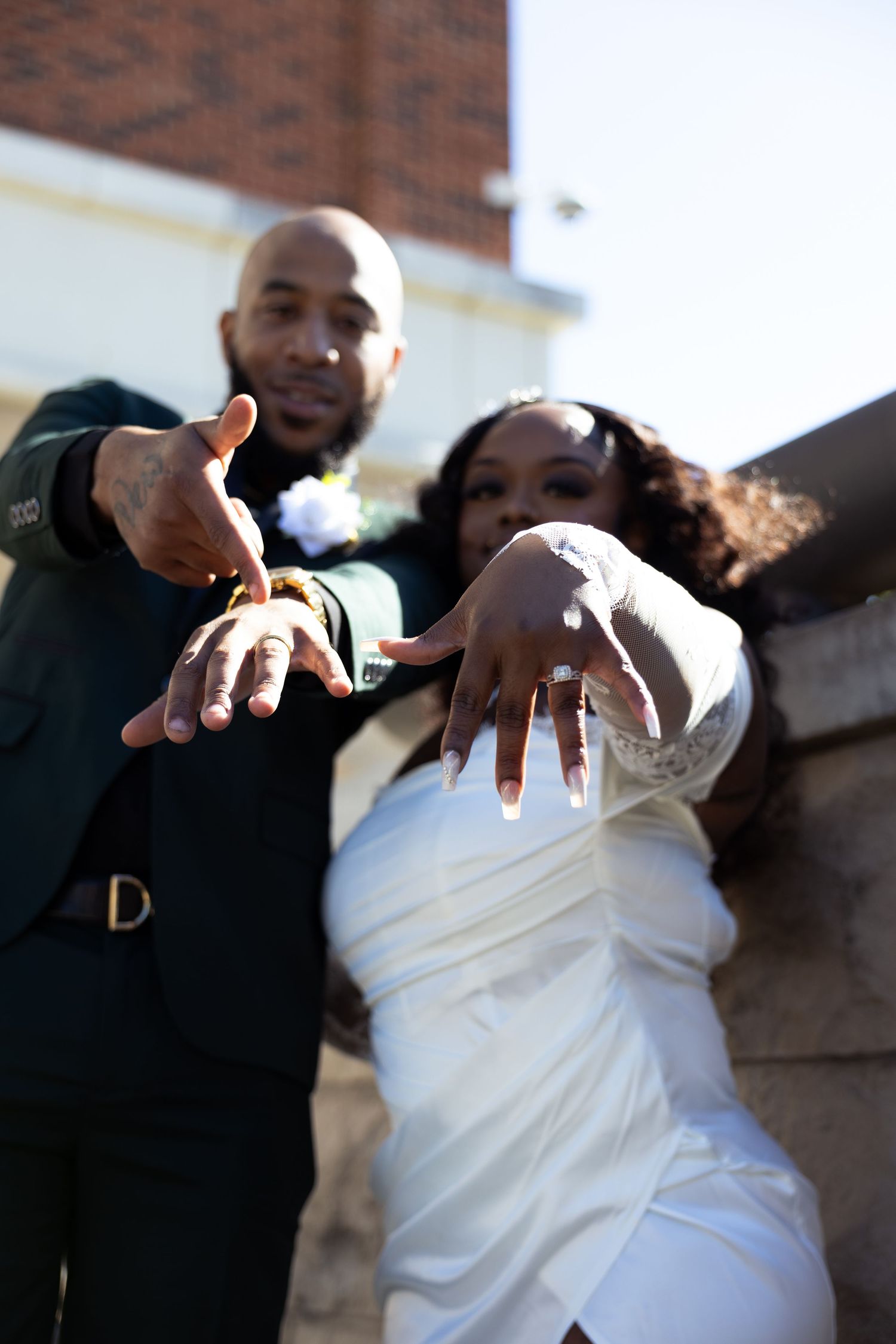 A newlywed couple proudly displays their wedding rings while posing outdoors in wedding attire.