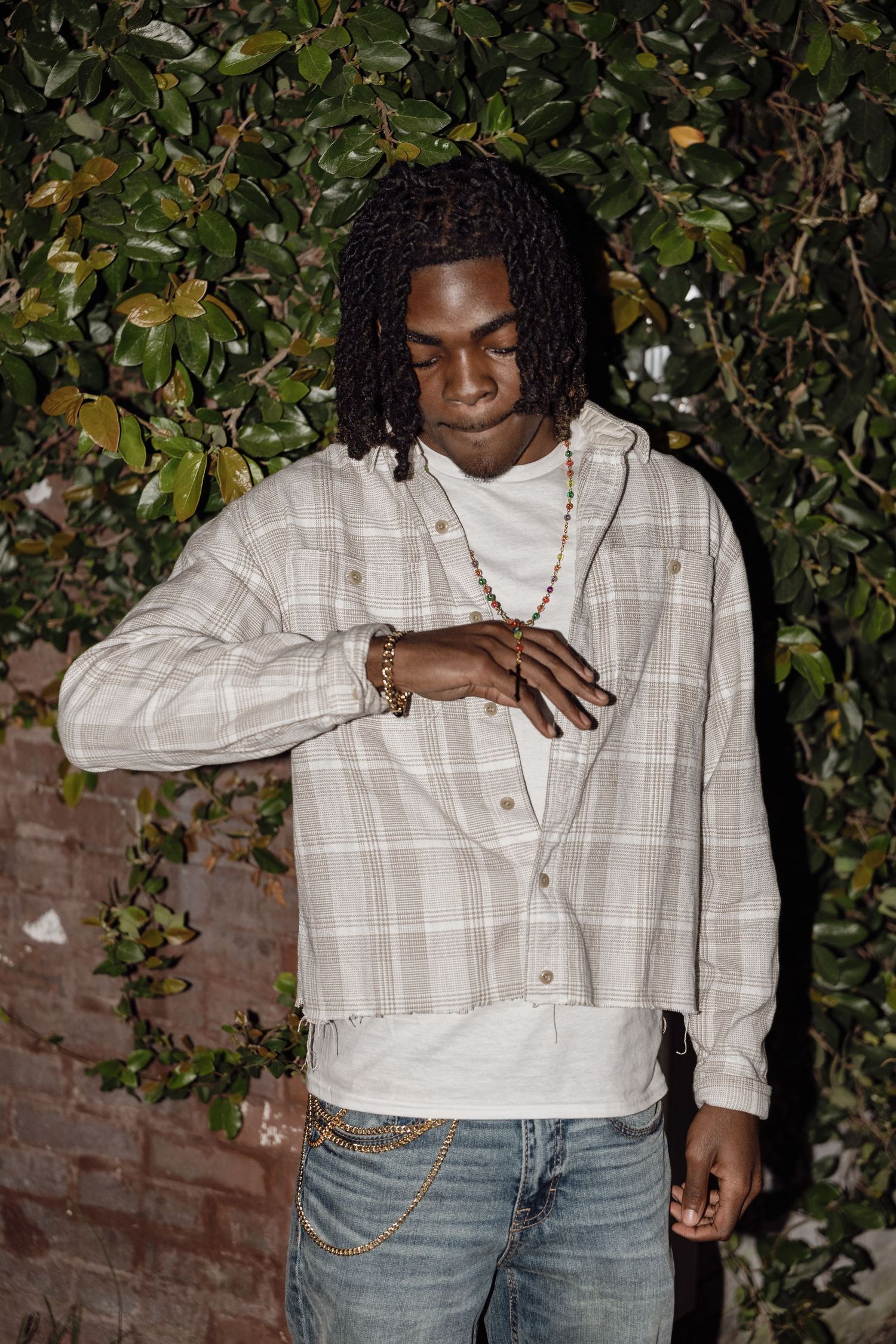 Young man with locs in plaid hoodie shirt and jeans poses outdoors near green foliage wall.