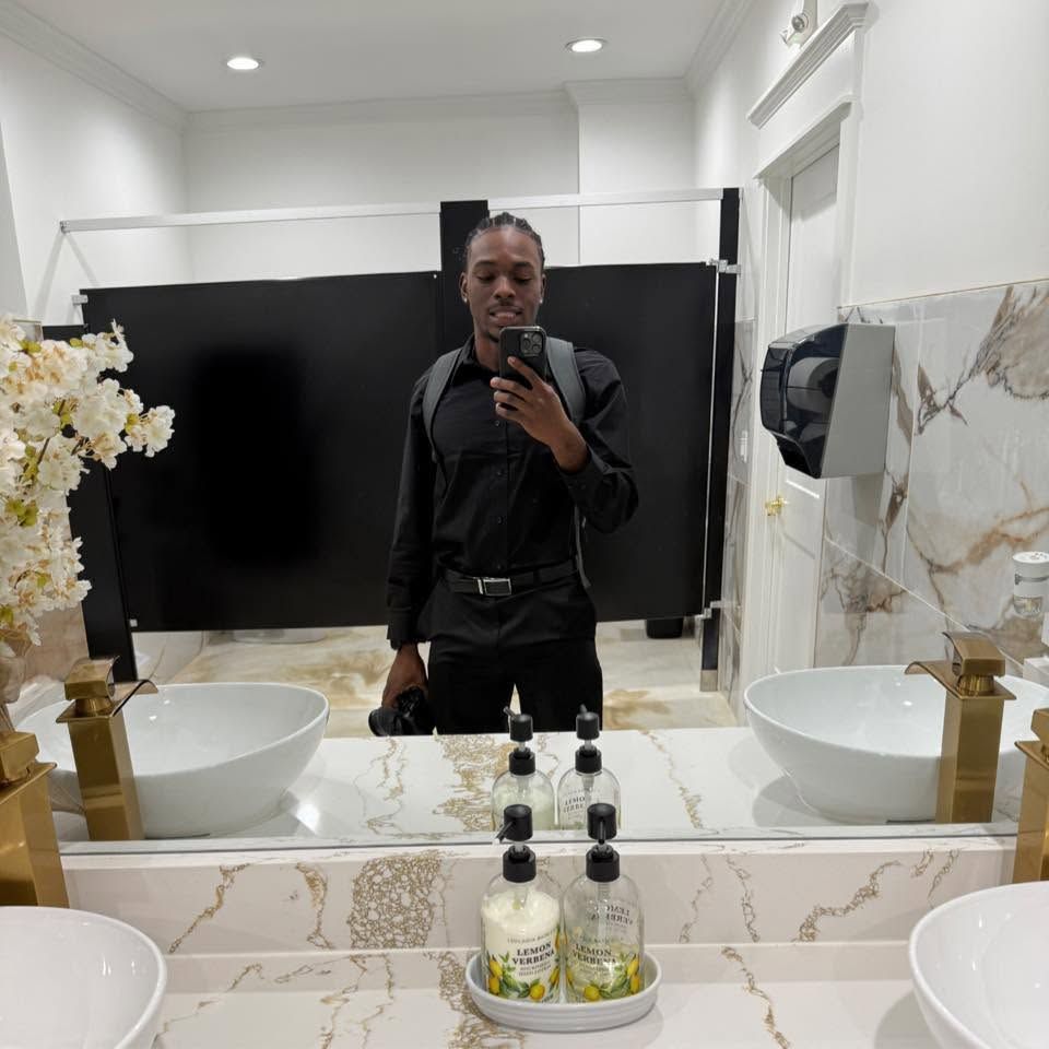 Man in all-black outfit takes mirror selfie in elegant marble bathroom with white vessel sinks.