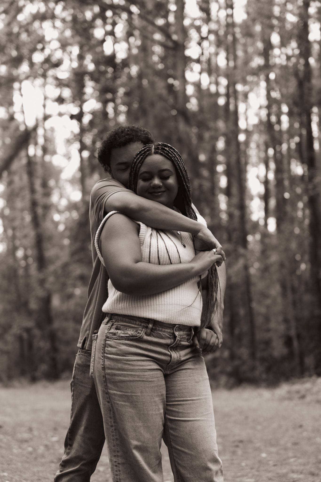 Black and white photo of couple embracing warmly in a forest setting among tall trees.