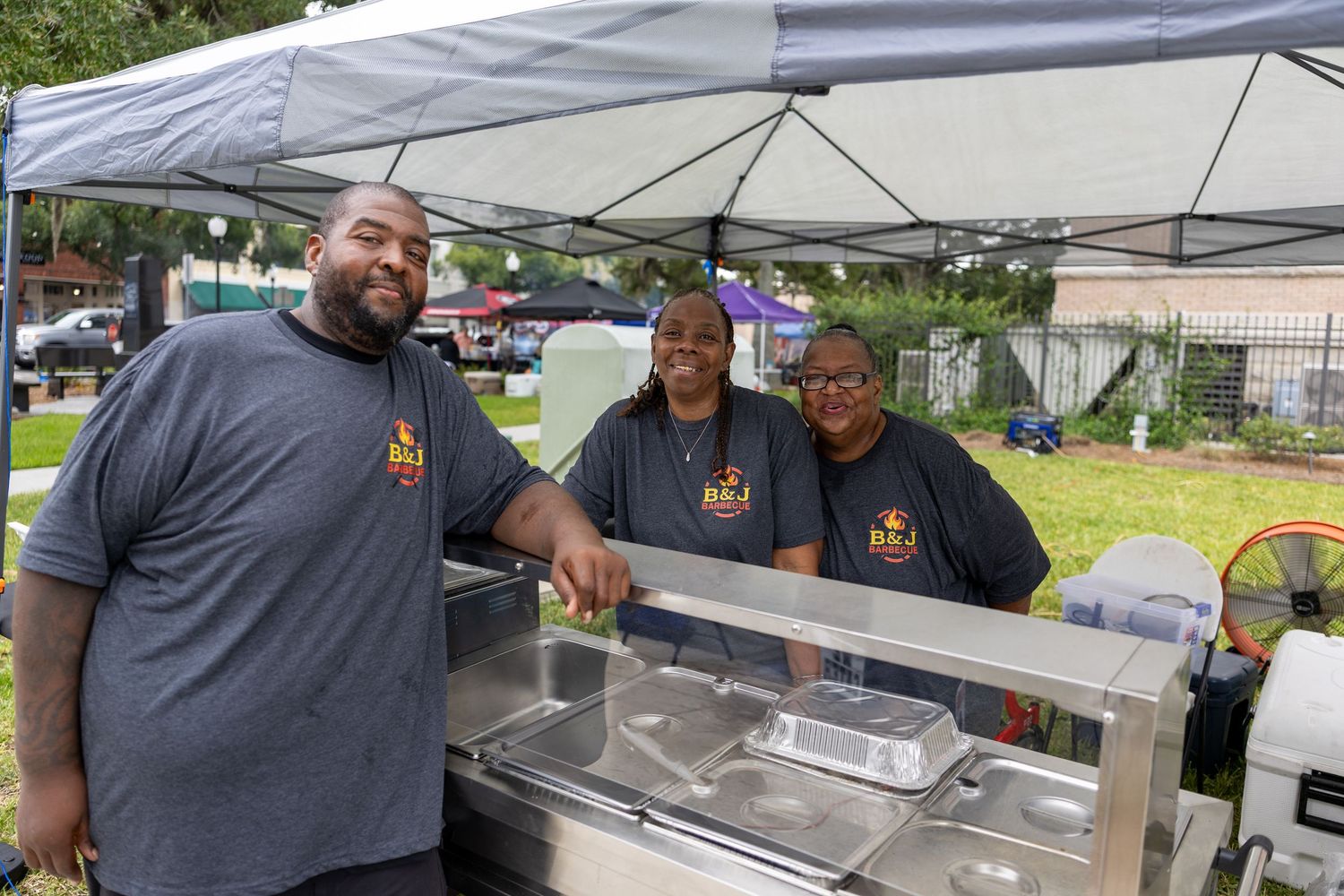 Three people in matching gray shirts standing behind food service equipment at an outdoor vendor booth.