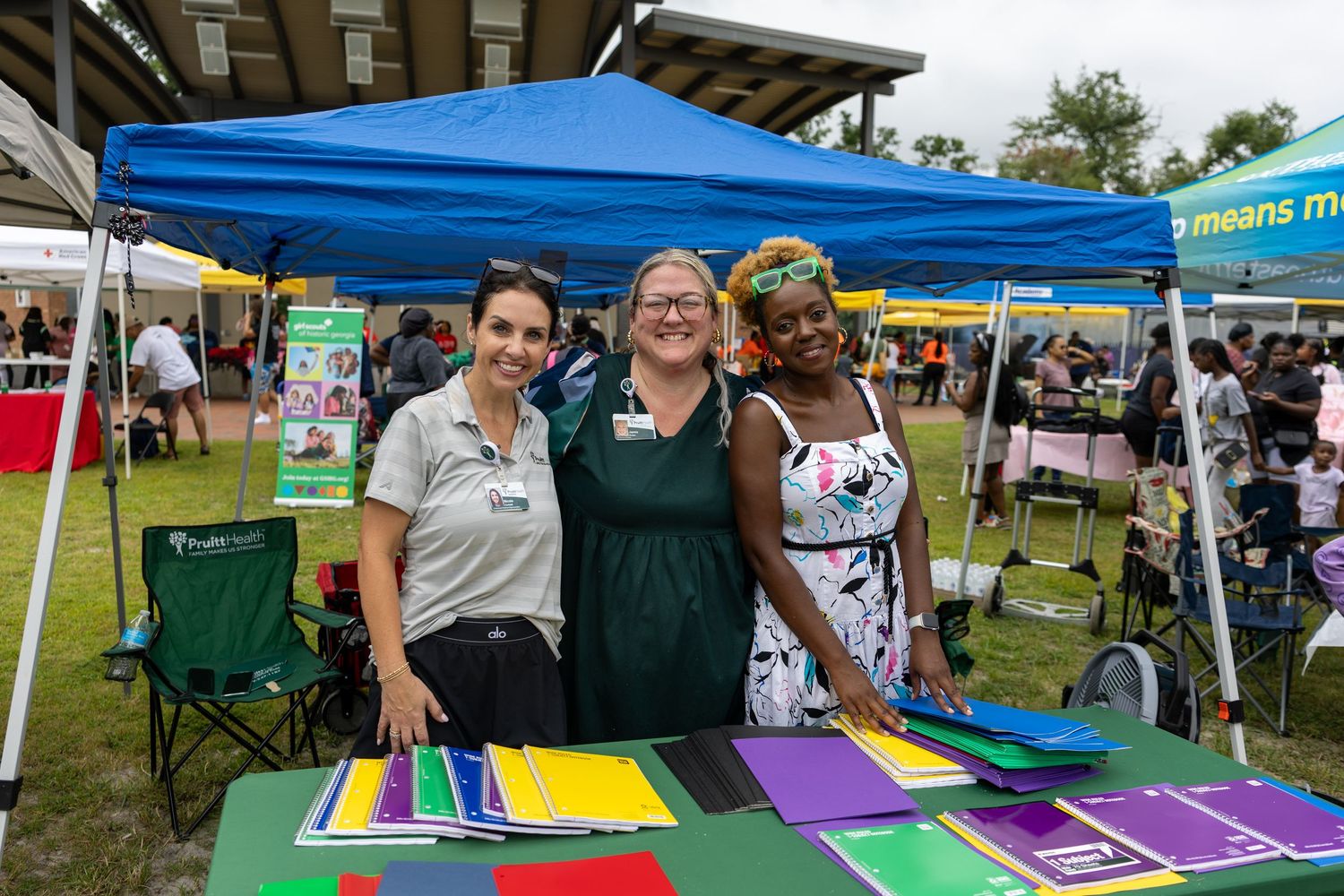 Three women smile together at outdoor community event booth with colorful materials on table.