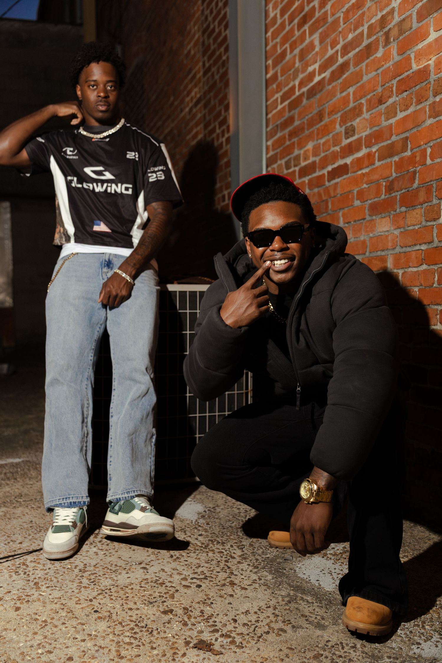 Two stylish young men pose against brick wall at night in urban streetwear outfits.