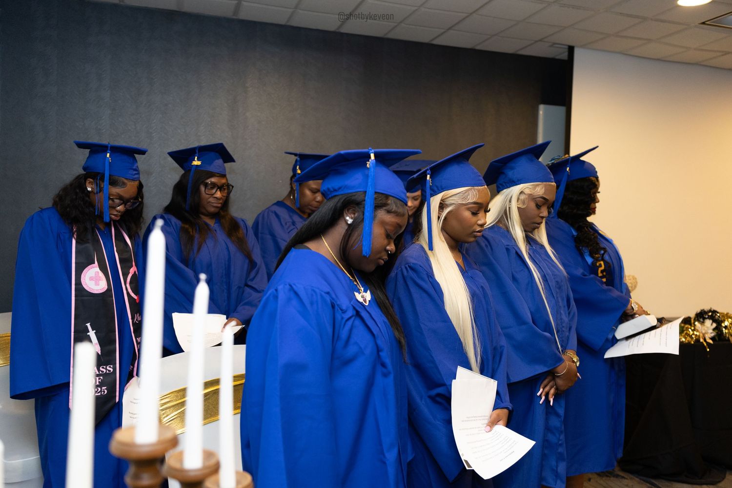 Group of graduates in blue caps and gowns standing during a graduation ceremony holding papers.