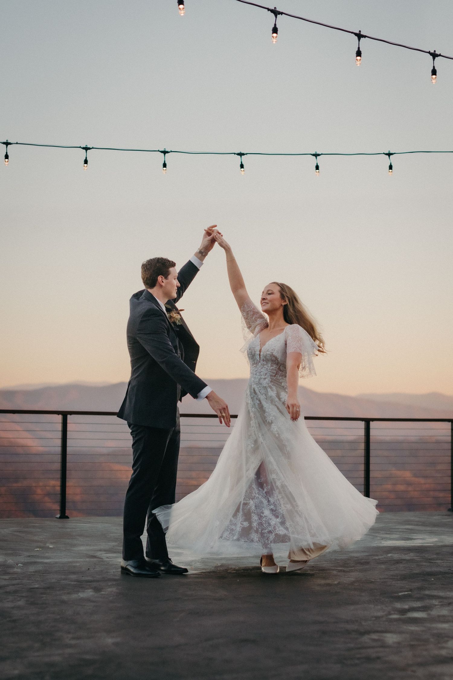 A couple dances together at sunset on a rooftop with string lights overhead.