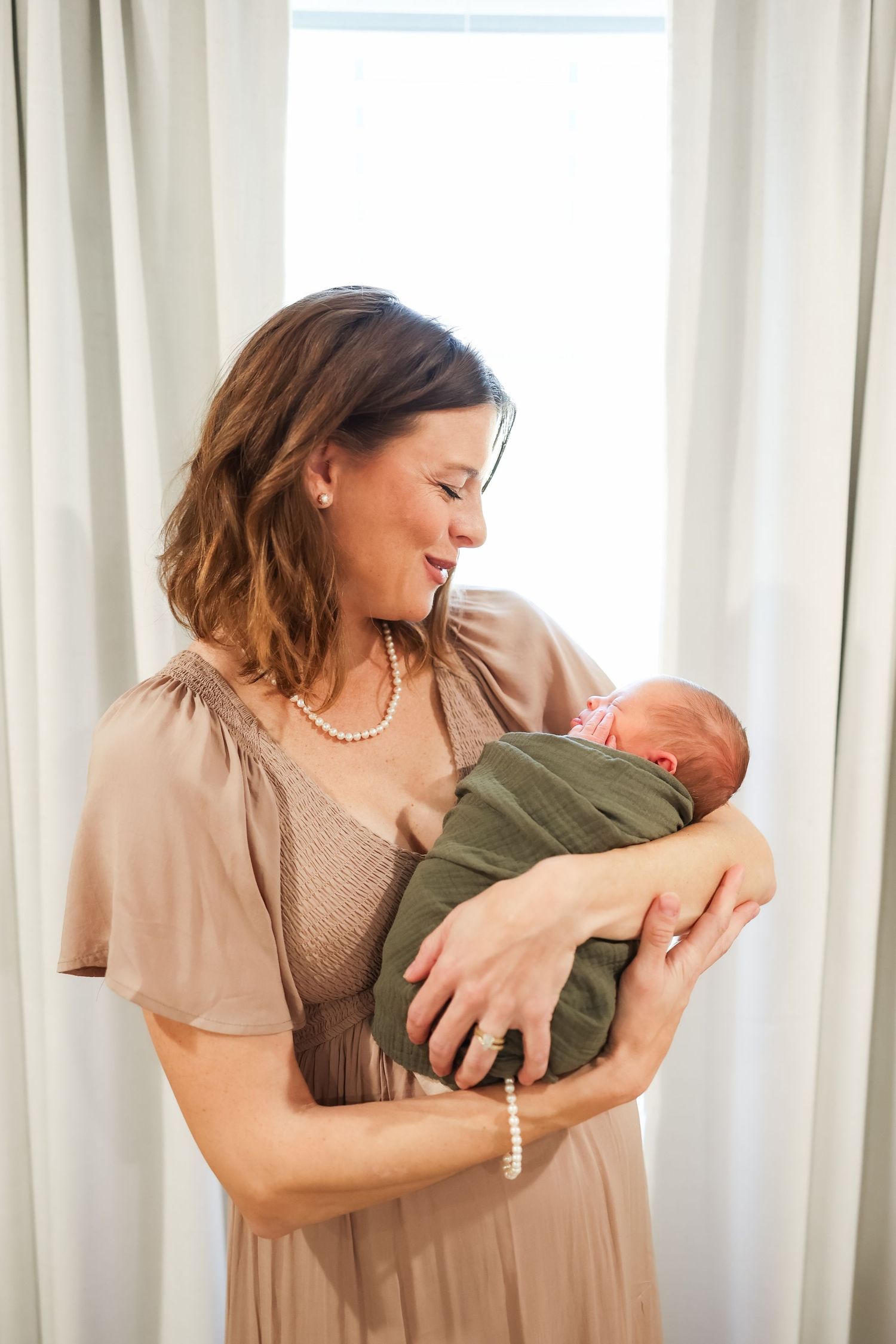 A tender moment in a sunlit nursery as a woman in a flowy dress cradles a newborn baby.