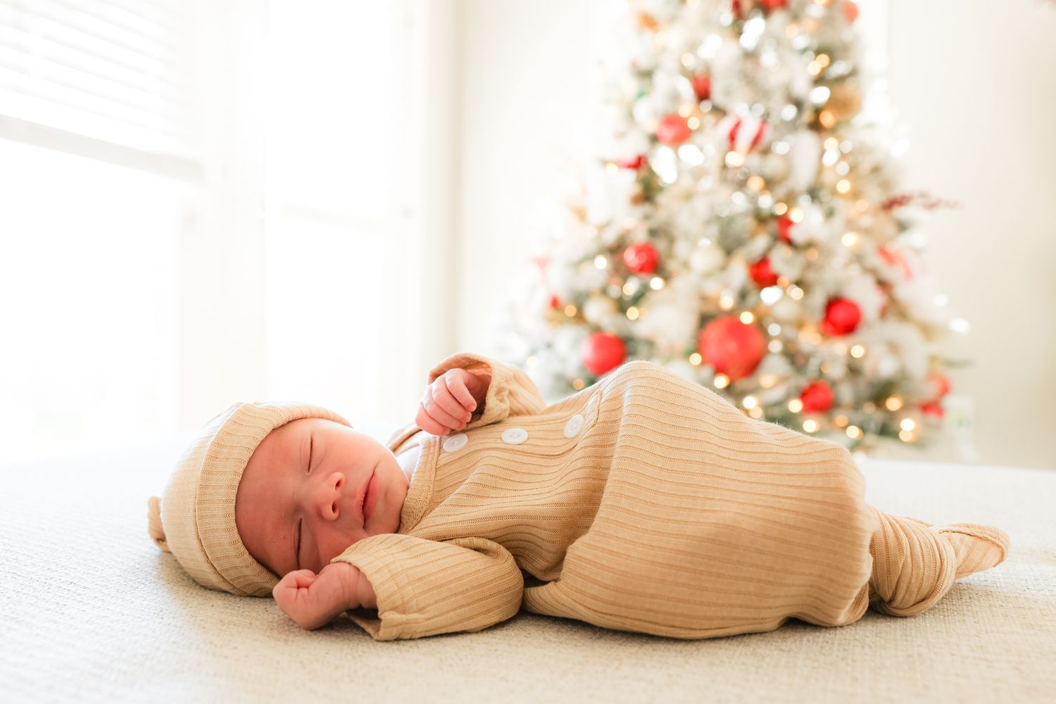 Sleeping newborn in cream knit outfit lies peacefully in front of decorated Christmas tree.