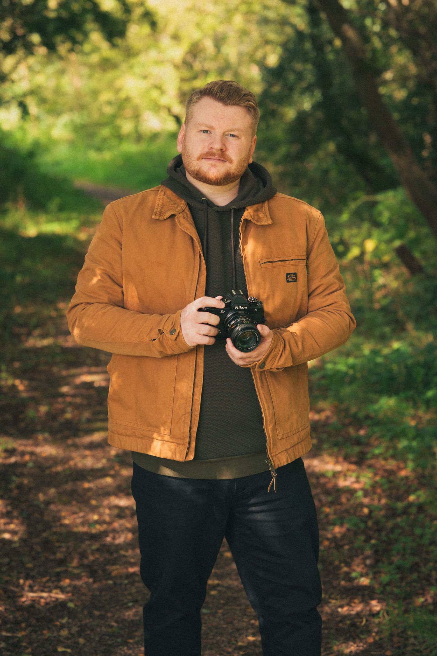 Male photographer holding a professional camera standing on a woodland path wearing an orange jacket.