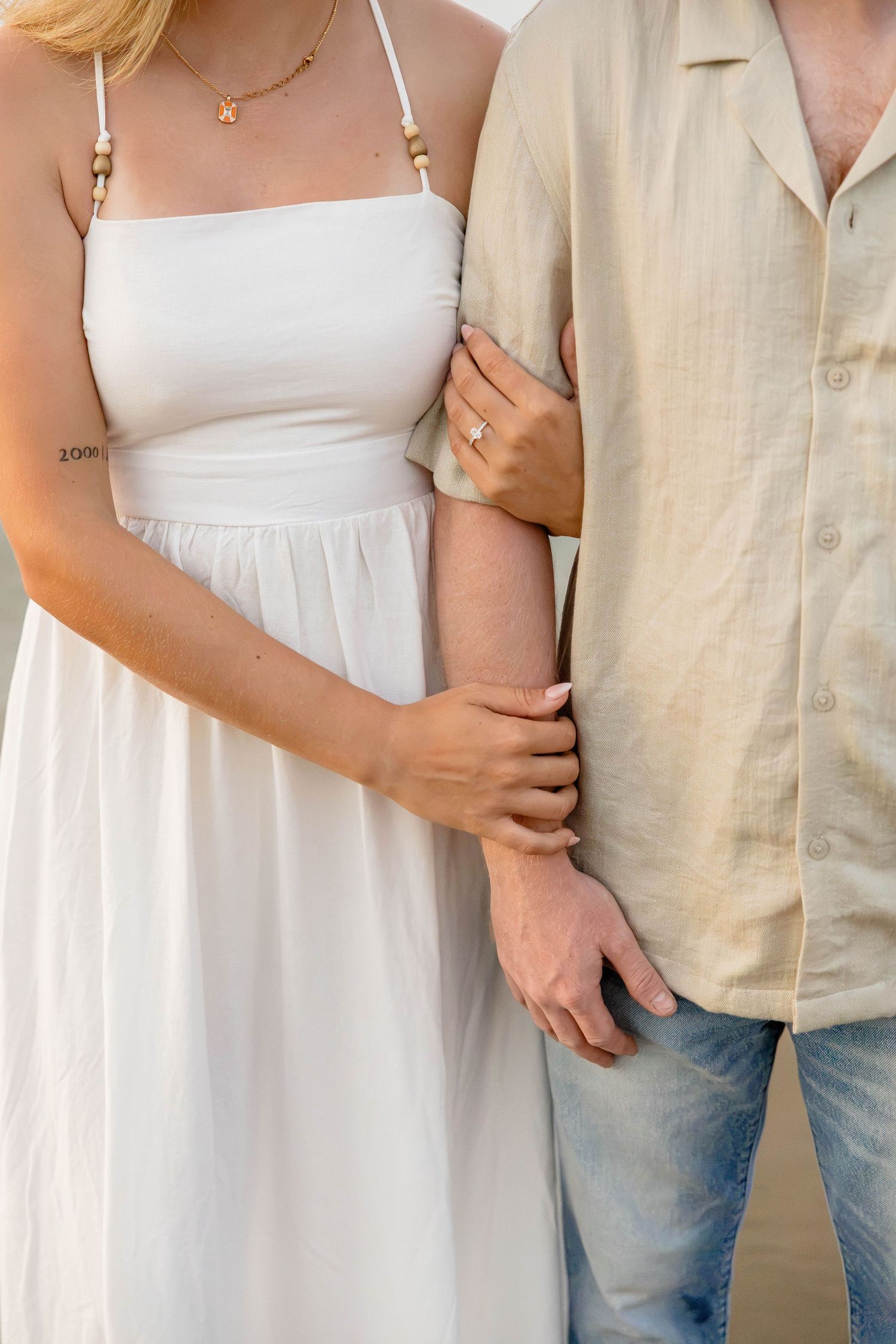 Couple in white dress and linen shirt holding arms, woman wearing engagement ring, beach setting.
