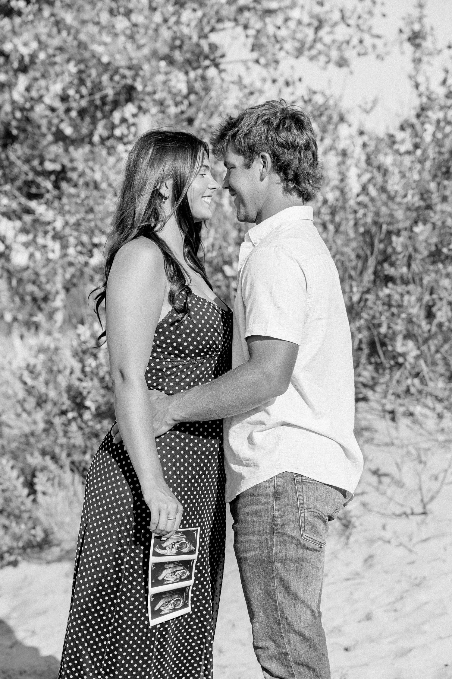 Black and white photo of a happy couple sharing an intimate moment, holding ultrasound photos, announcing their pregnancy outdoors.