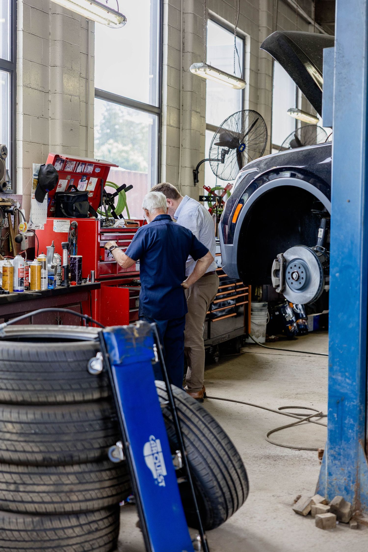Two mechanics inspect a lifted car in a professional auto repair shop with tires and tools visible.