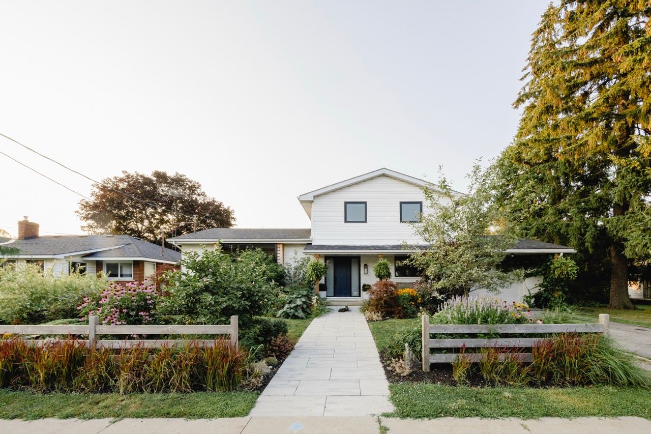 White two-story suburban home with a charming front garden, wooden fence, and tree-lined pathway at dusk.