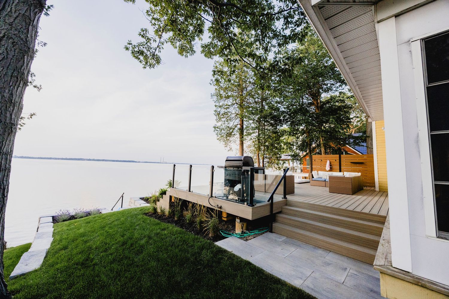 Waterfront property deck with outdoor grill, lush lawn, and calm lake views surrounded by mature trees.
