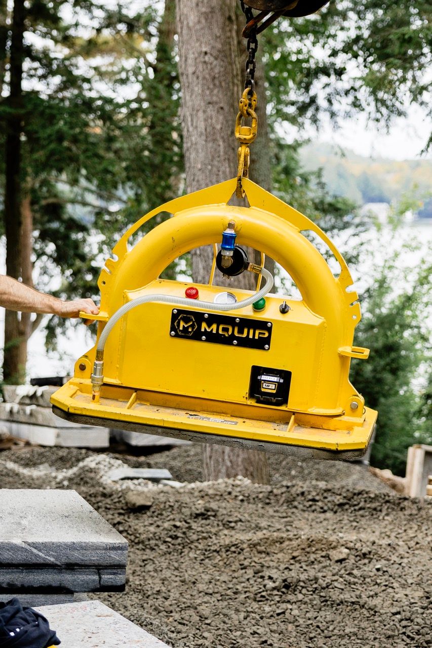 Yellow MQUIP vacuum stone lifting device hanging from excavator arm at outdoor lakeside landscaping site.