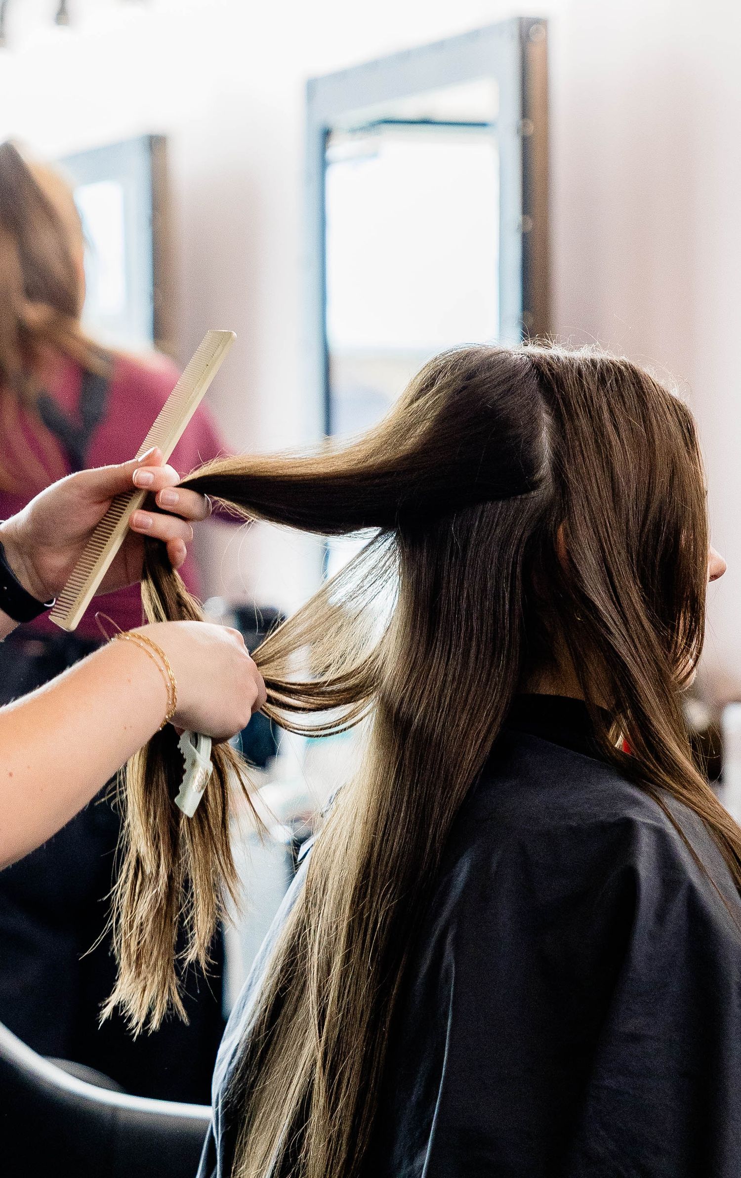 Hairstylist combing long brunette hair with highlights at a professional salon, mirror visible in background.