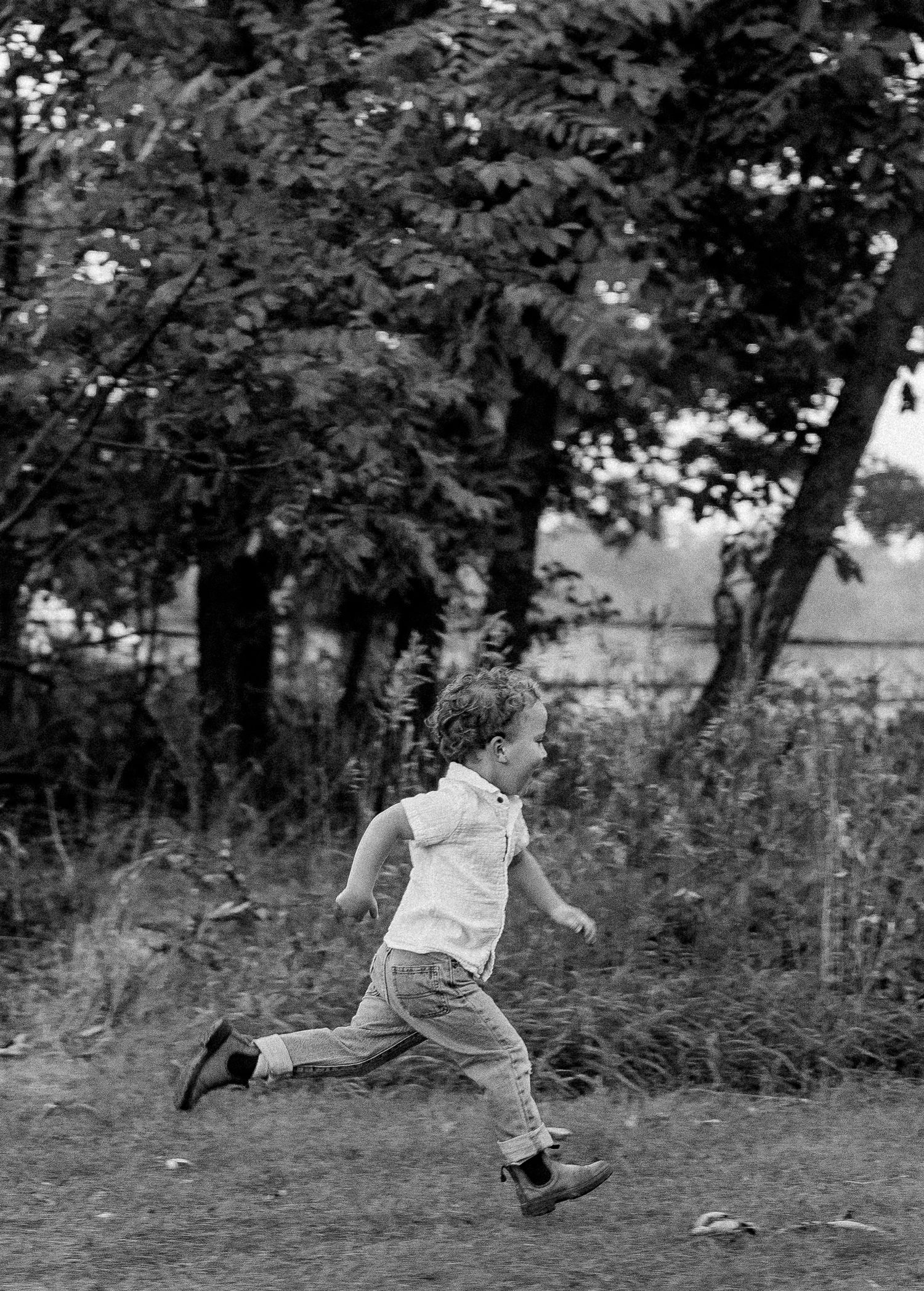 Black and white photo of a young child running joyfully through a grassy field with trees in the background.