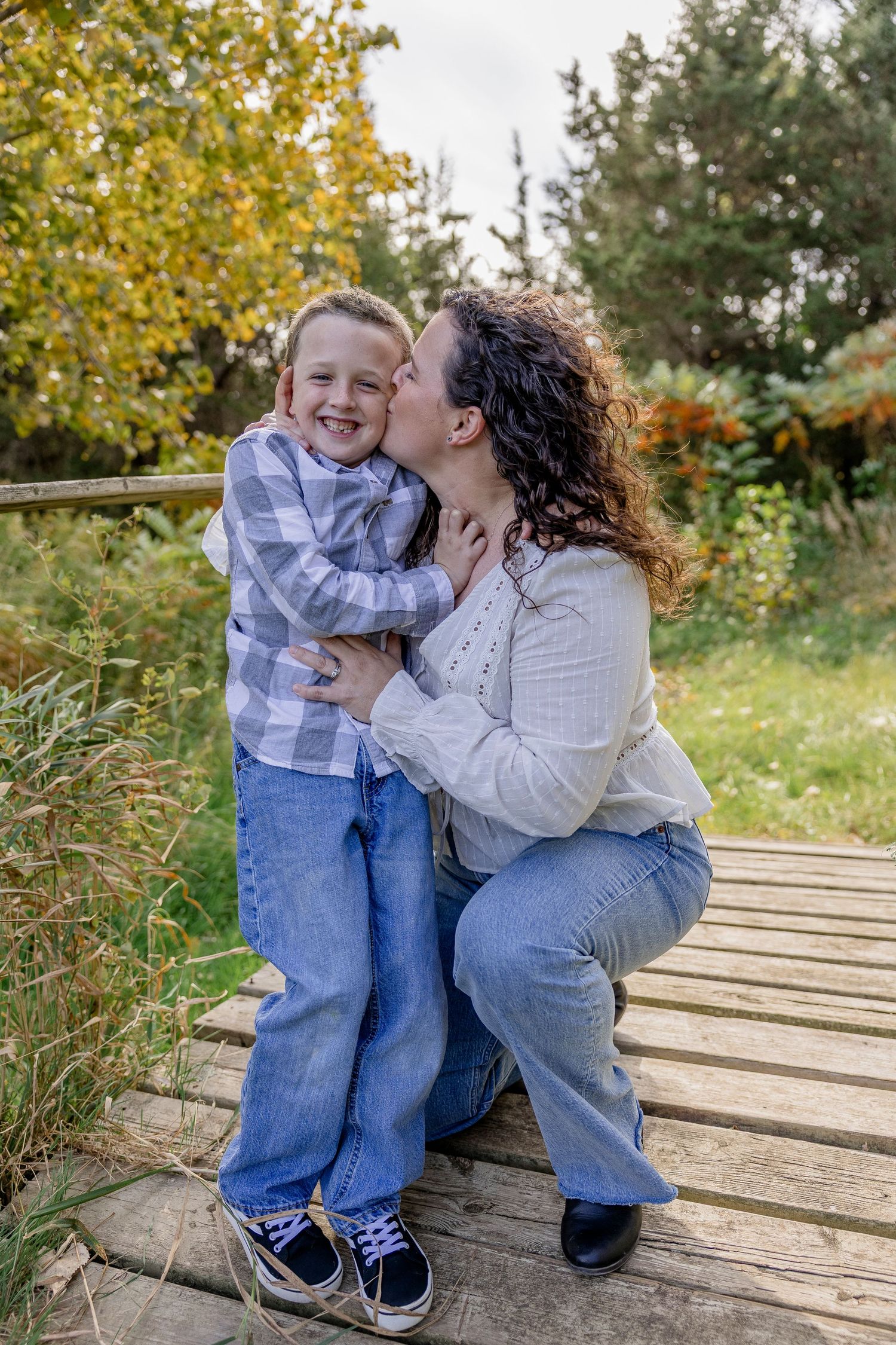 A curly-haired mother in white kisses her laughing young son on a wooden path amid lush autumn greenery.