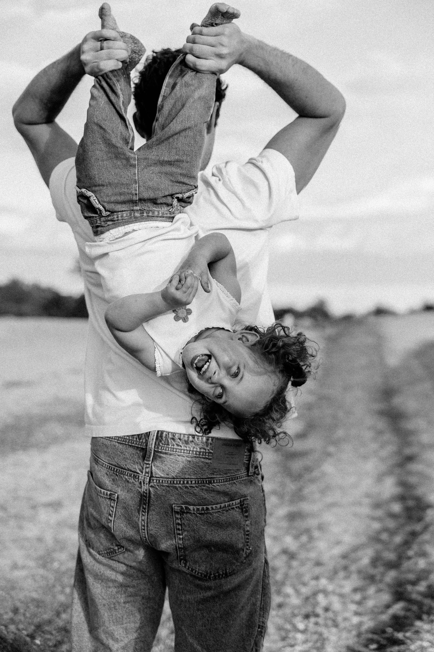 Black and white photo of parent playfully holding toddler upside down outdoors in a field.
