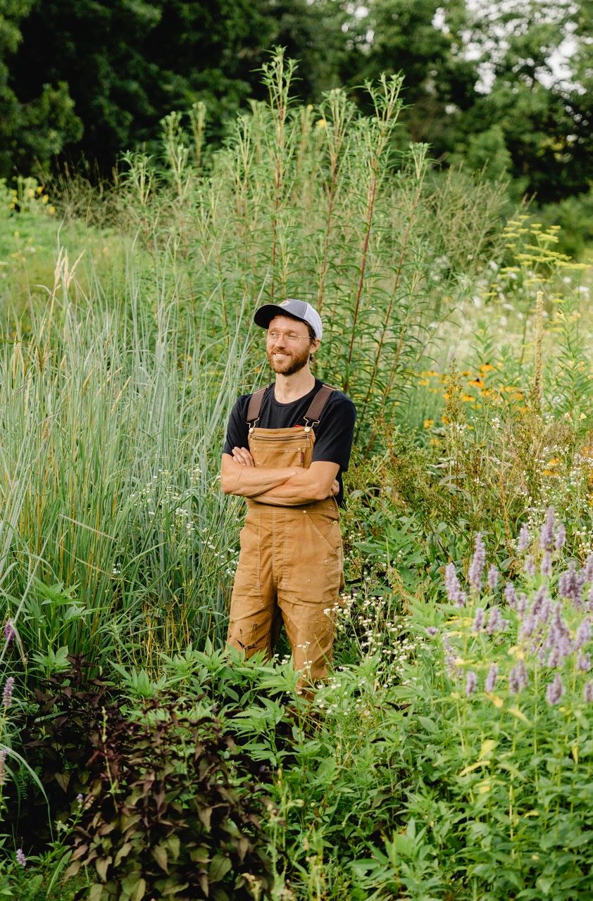 Man in overalls and cap standing with arms crossed in lush garden.