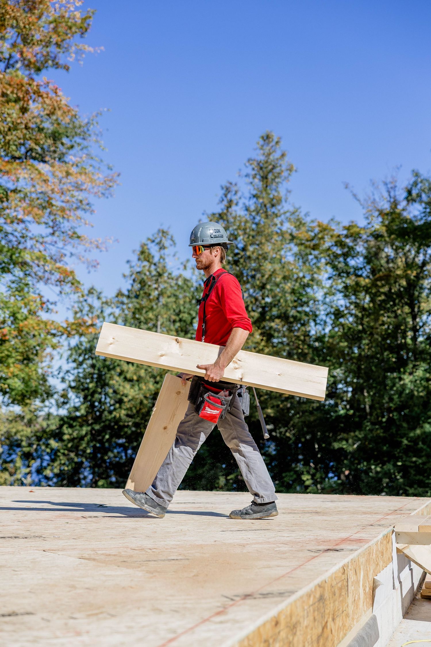 Construction worker in red shirt and hard hat carrying a wooden plank on a building site under blue sky.