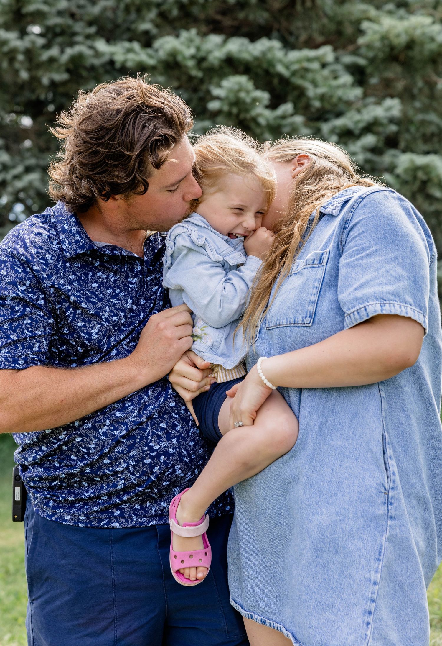 A happy family with a man and woman kissing their laughing toddler girl outdoors near evergreen trees.