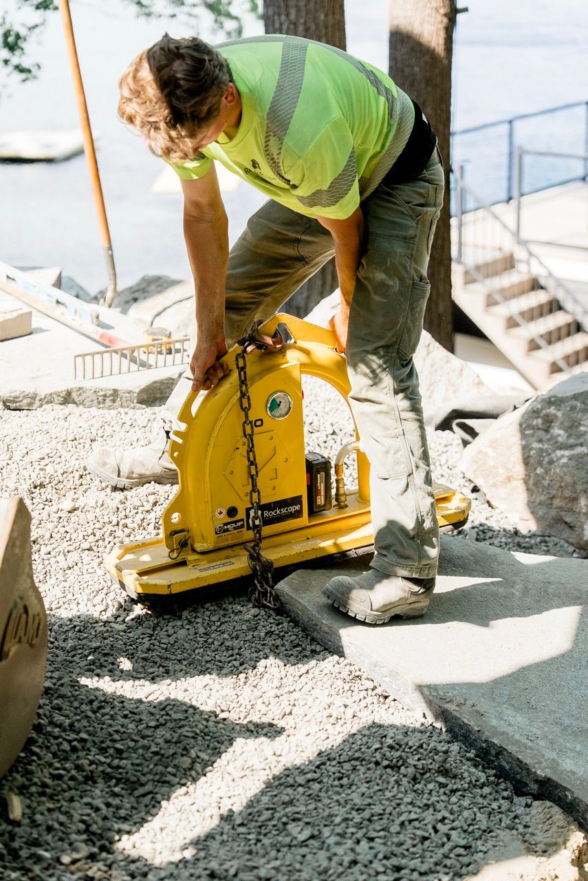 Landscaping worker in hi-vis vest operating yellow MQUIP vacuum stone lifter on dark gravel surface.