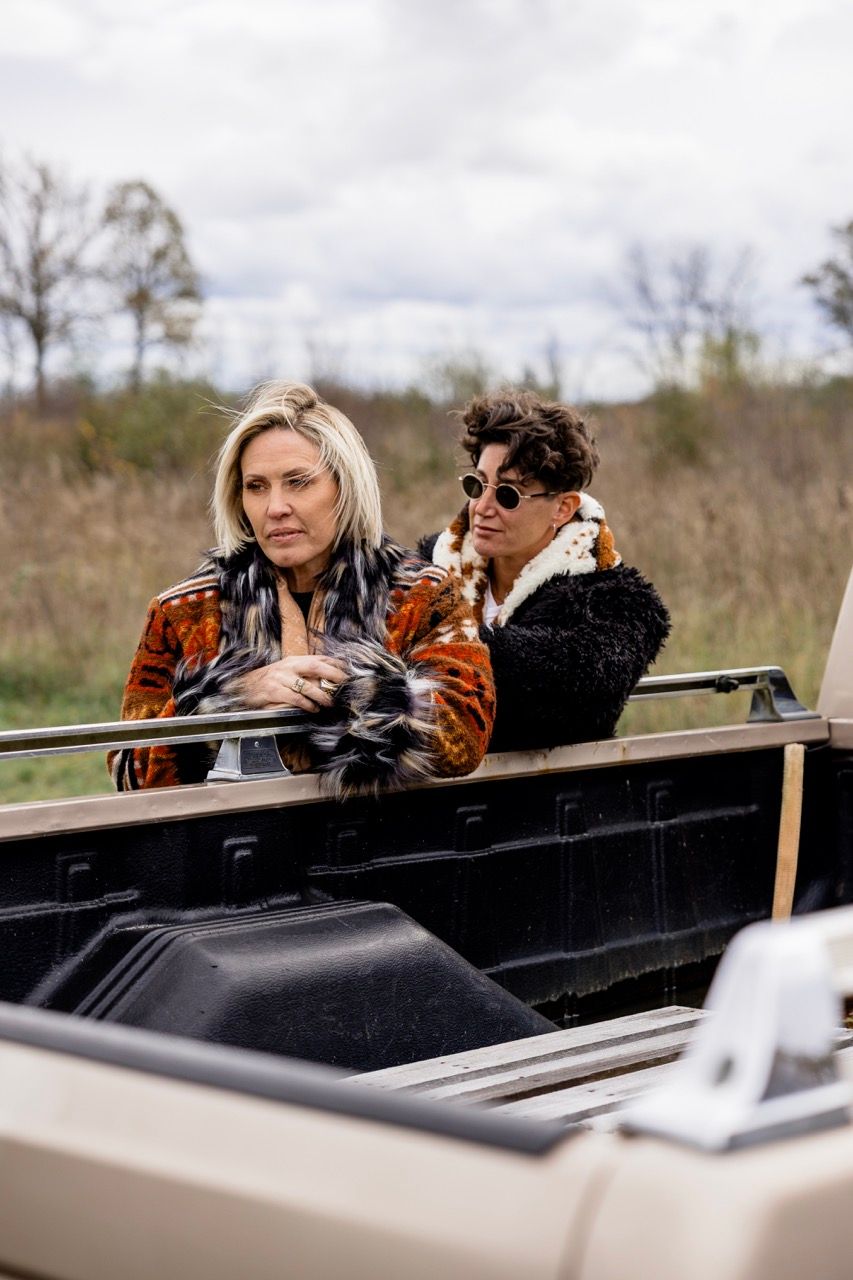 Two stylish women sitting in the bed of a vintage pickup truck in a rural autumn setting.
