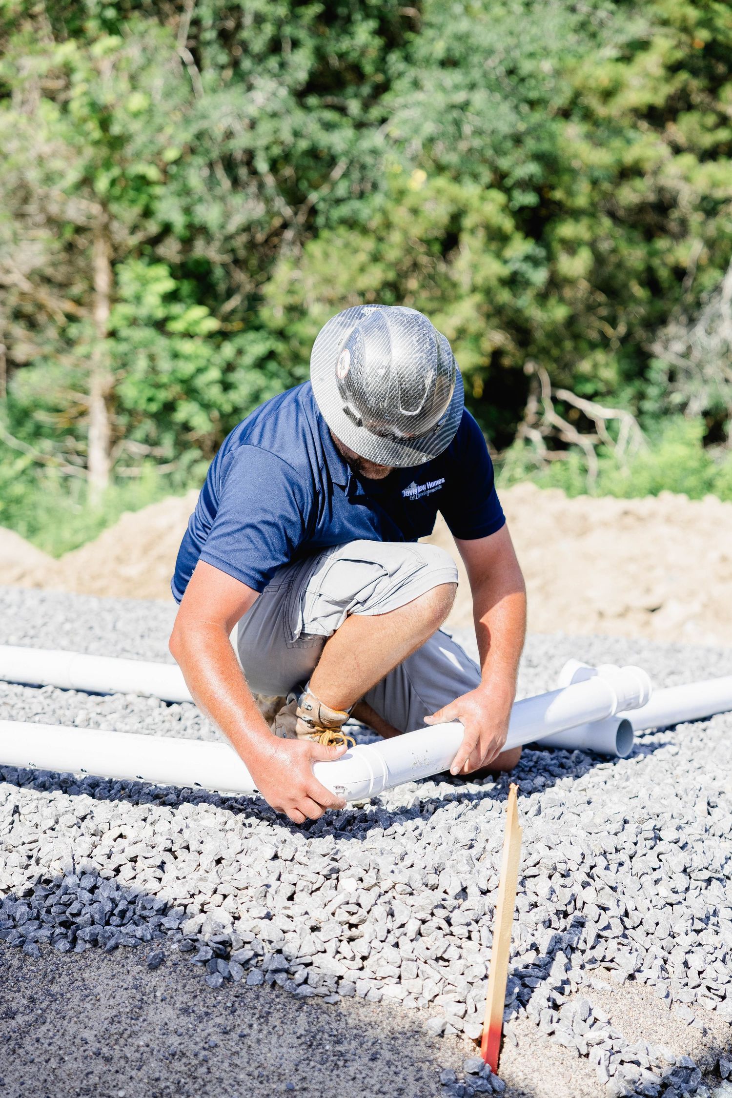 Construction worker in gray hard hat crouching on gravel installing white PVC pipes at a building site.