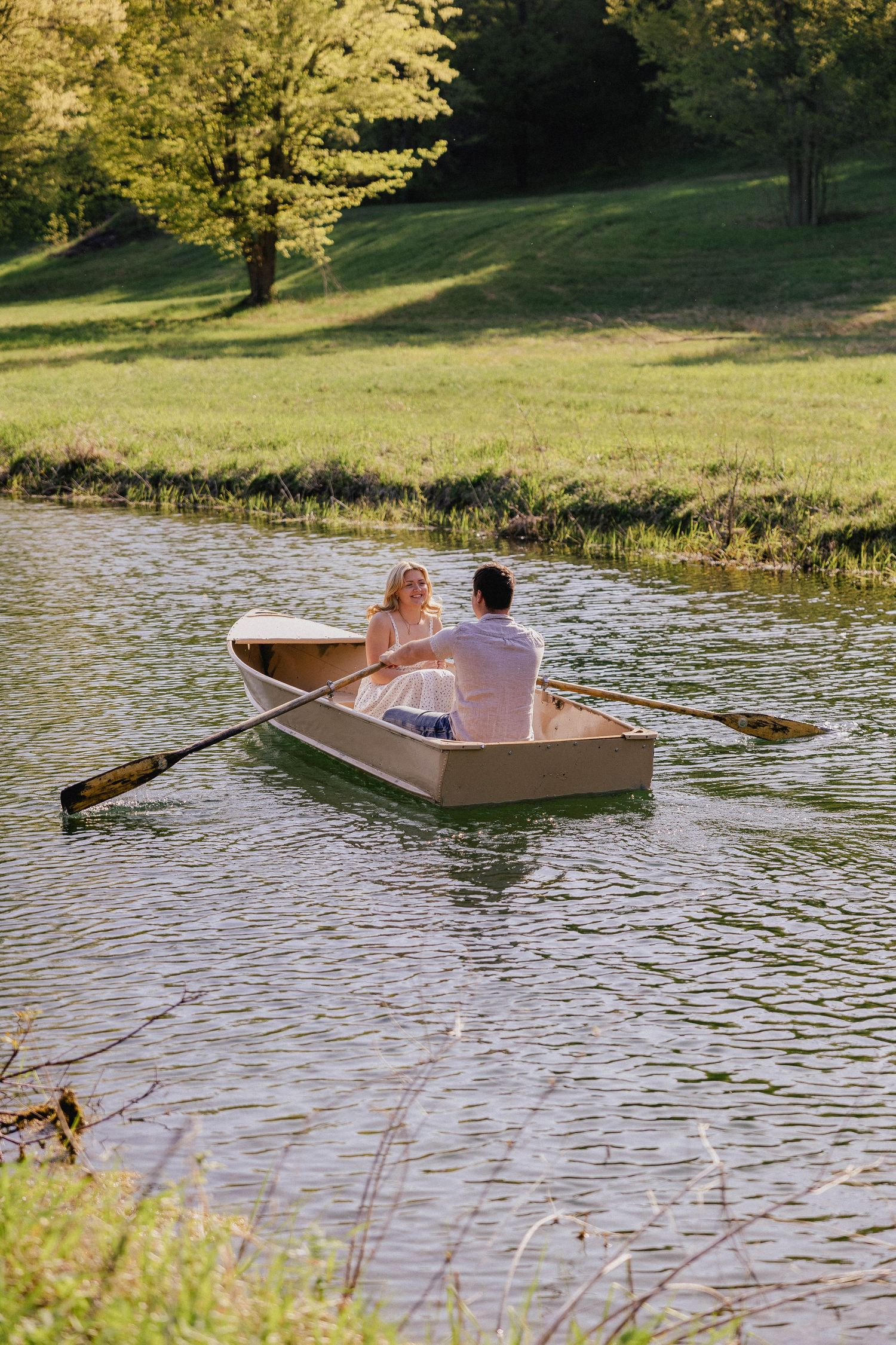 Couple rowing a wooden boat on a calm green lake surrounded by lush trees and meadows.