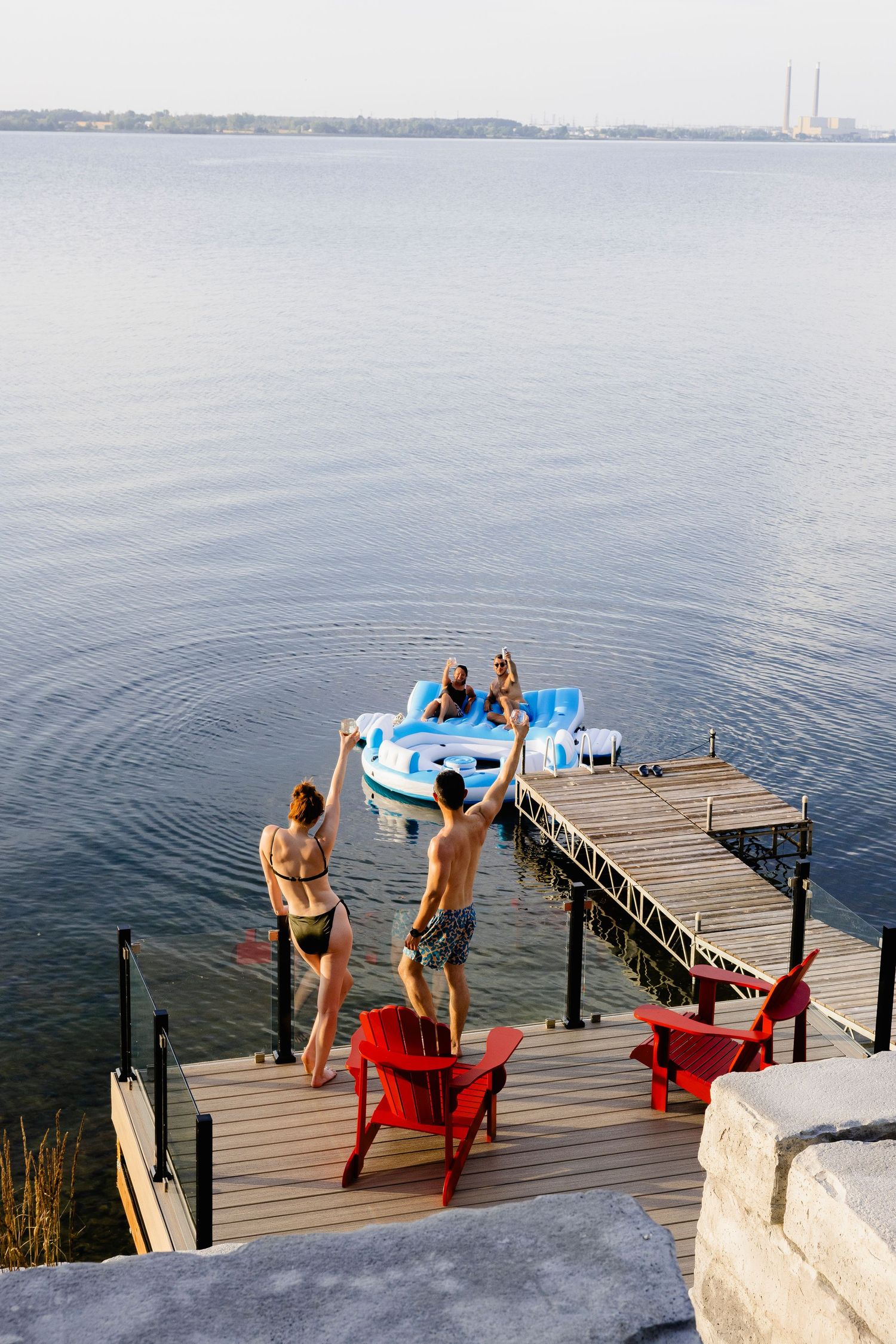 Friends on a wooden dock wave to people on a blue pedal boat on a calm lake on a sunny day.