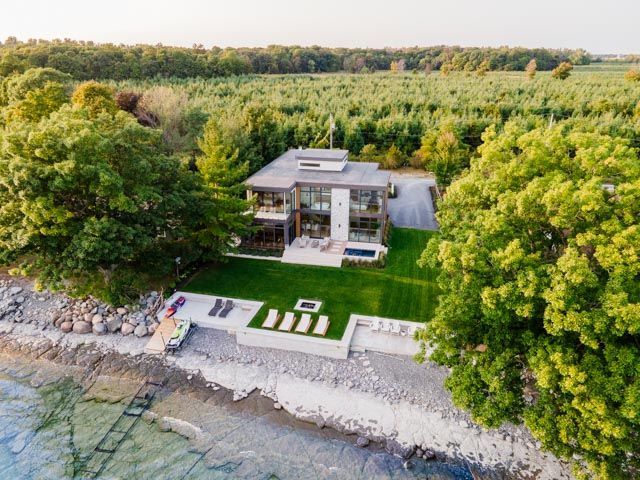 Aerial view of a modern waterfront home surrounded by lush trees with a manicured lawn and rocky shoreline.