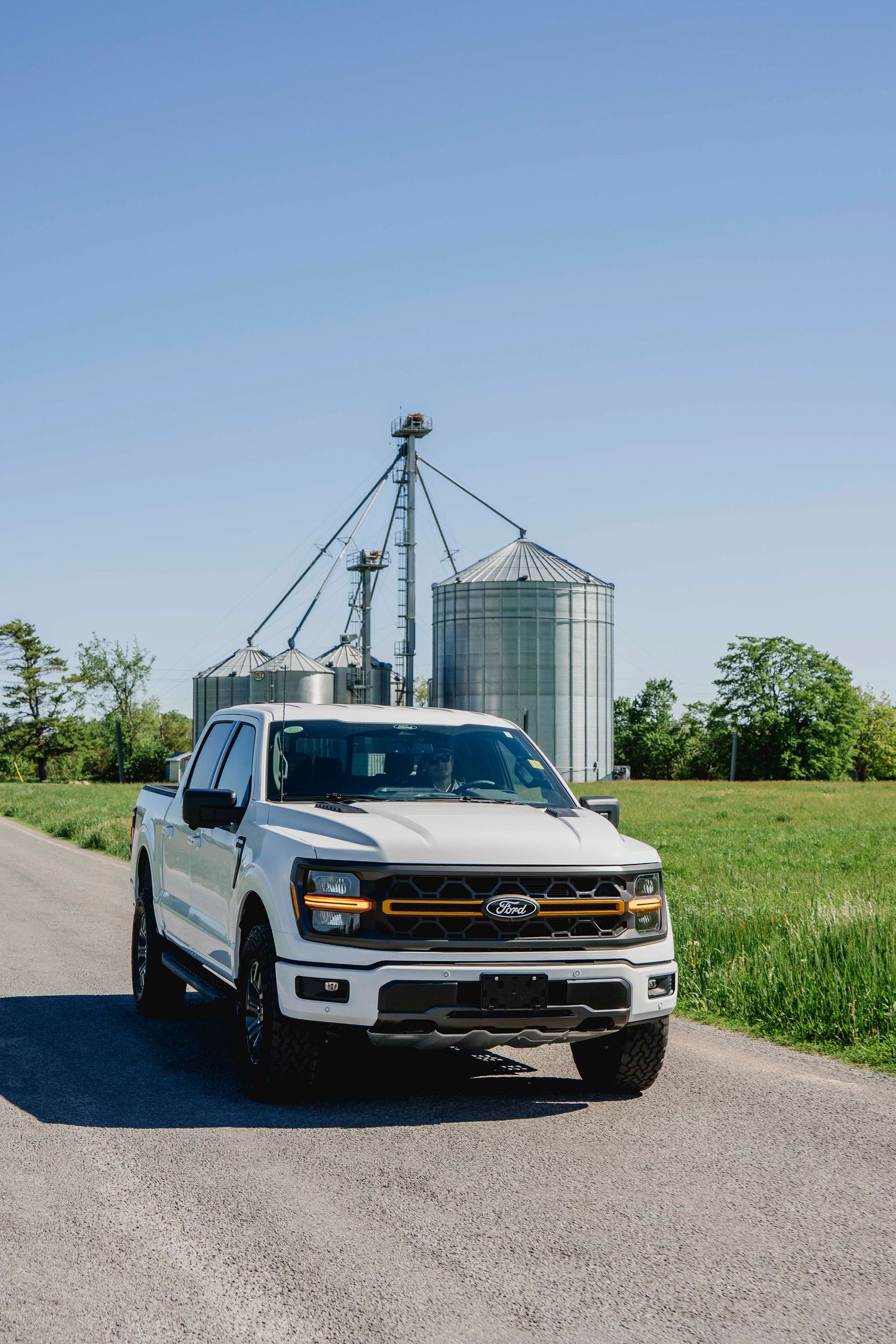 White Ford F-150 truck on rural road with grain silos and green farmland in background.