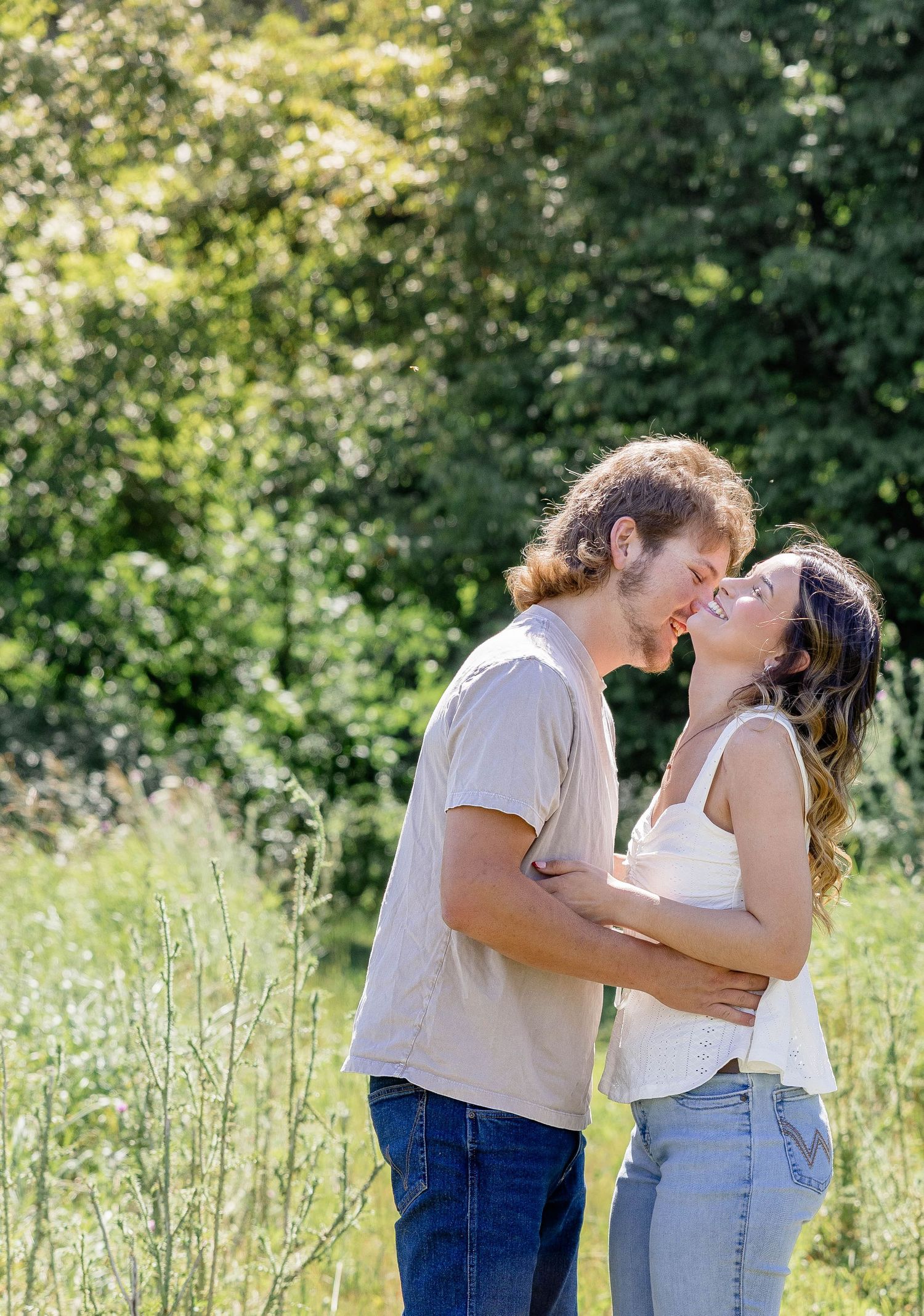 A couple shares a romantic almost-kiss while embracing in a sunny green meadow, both wearing casual jeans and tops.