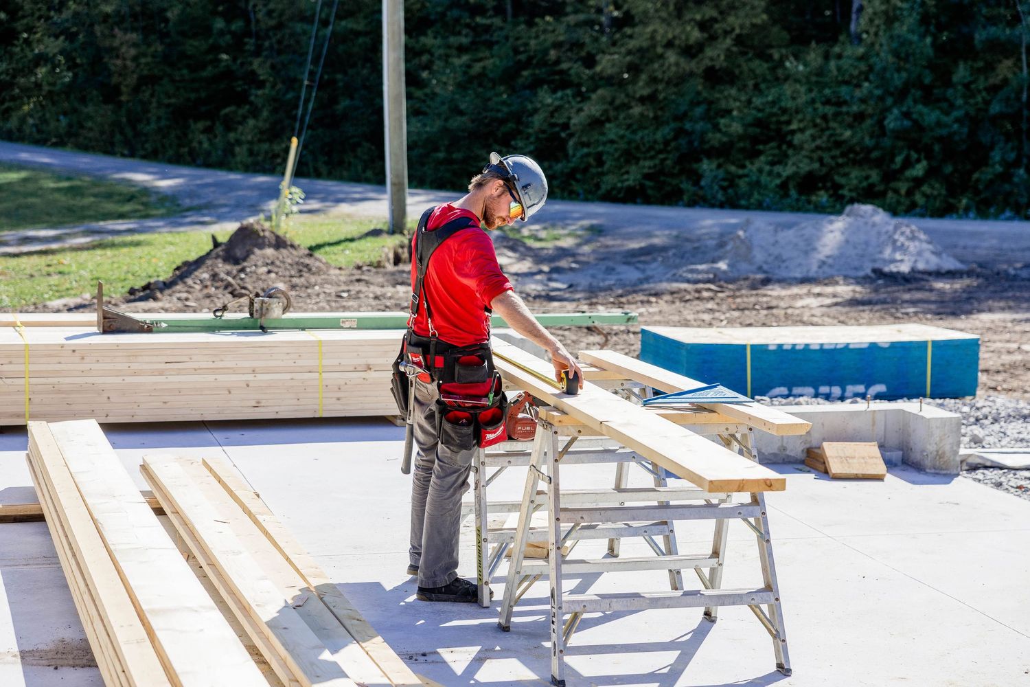 Construction worker in red shirt and safety gear measuring lumber at a building site on a sunny day.