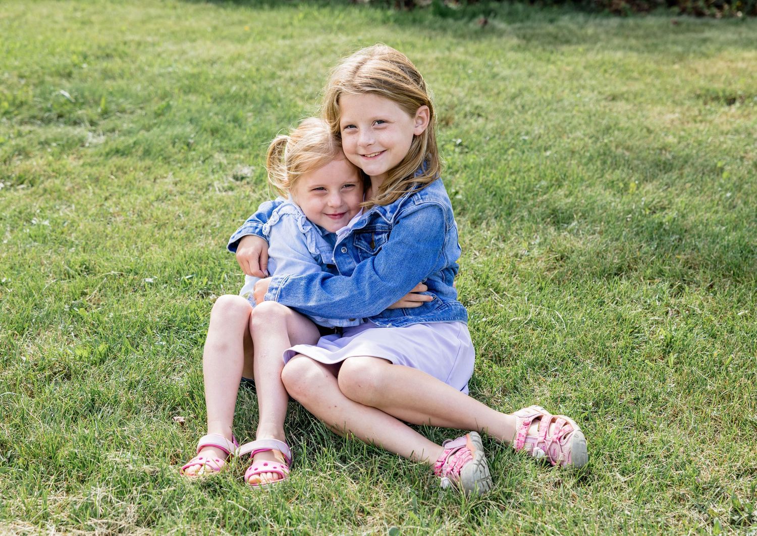 Two young girls in blue outfits hugging and smiling while sitting on green grass outdoors.