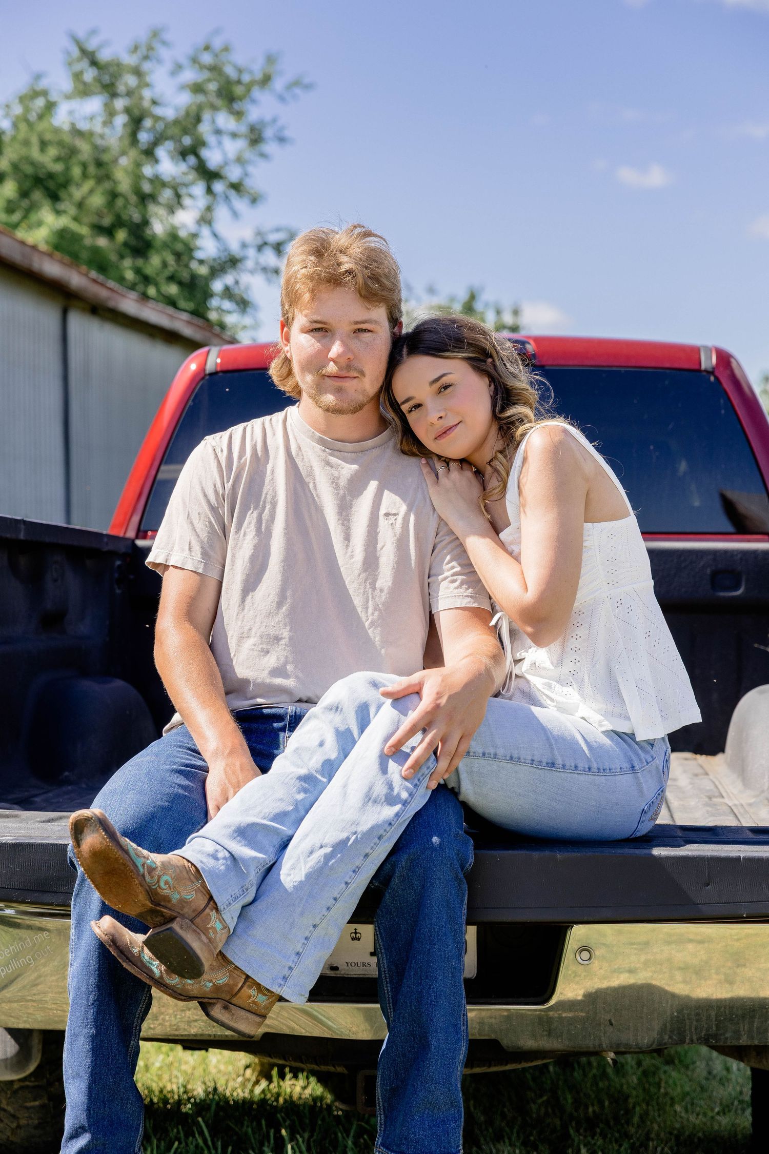 Young couple sitting together on a red pickup truck tailgate outdoors on a sunny day.