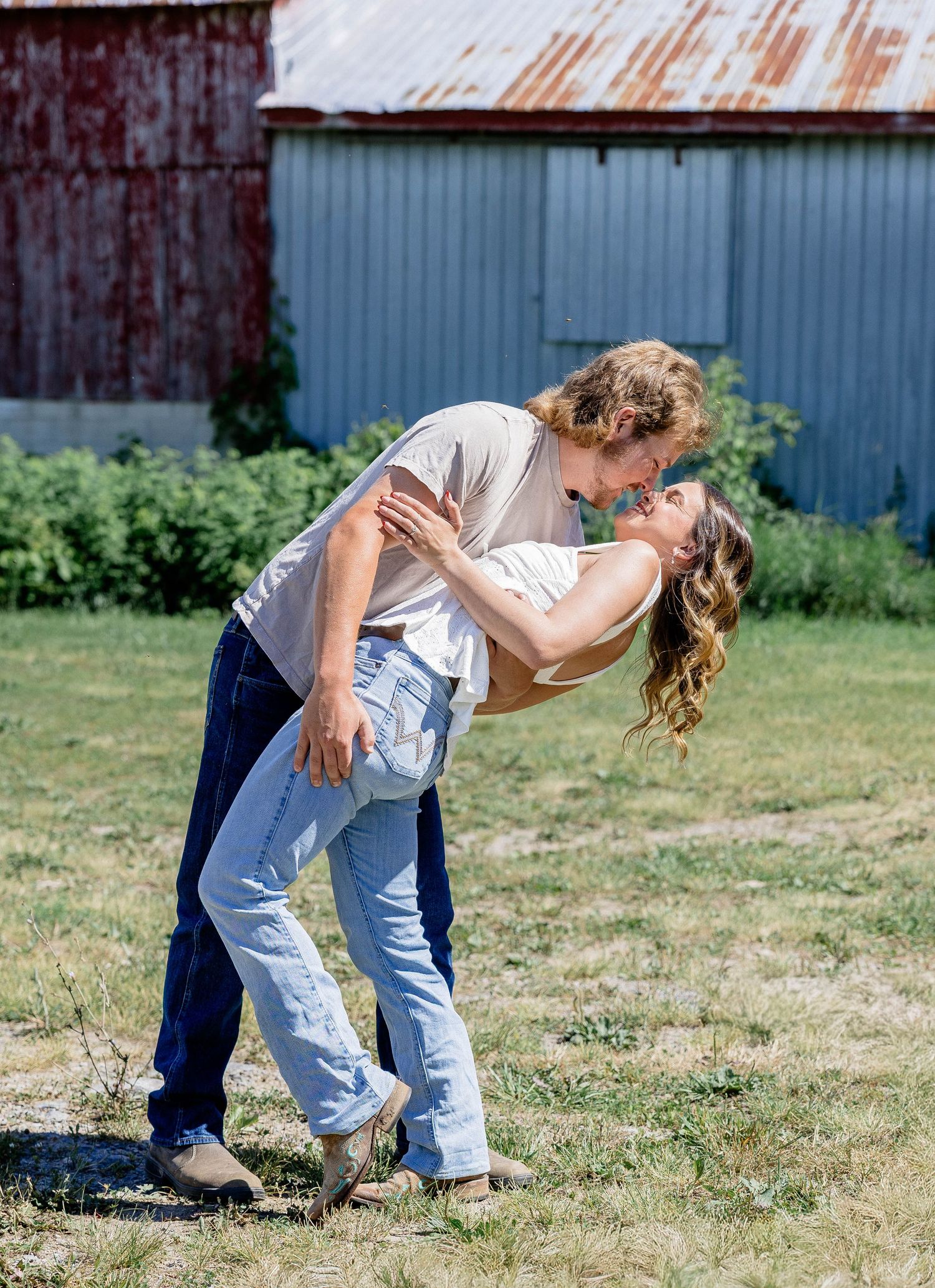 Couple sharing a romantic dip kiss outdoors near a rustic barn on a sunny day.