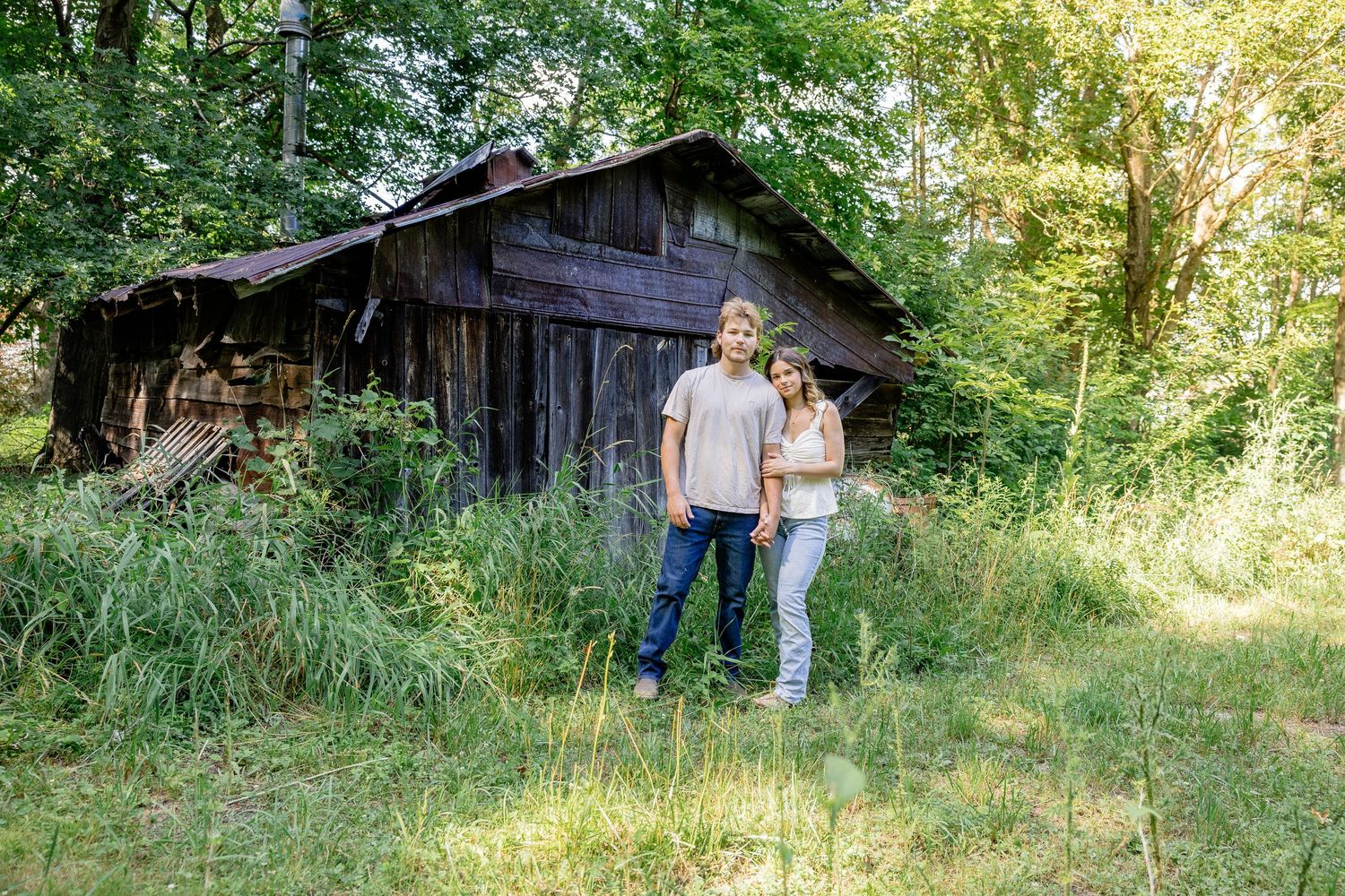 Couple standing together in front of a rustic wooden barn surrounded by lush green trees and tall grass.