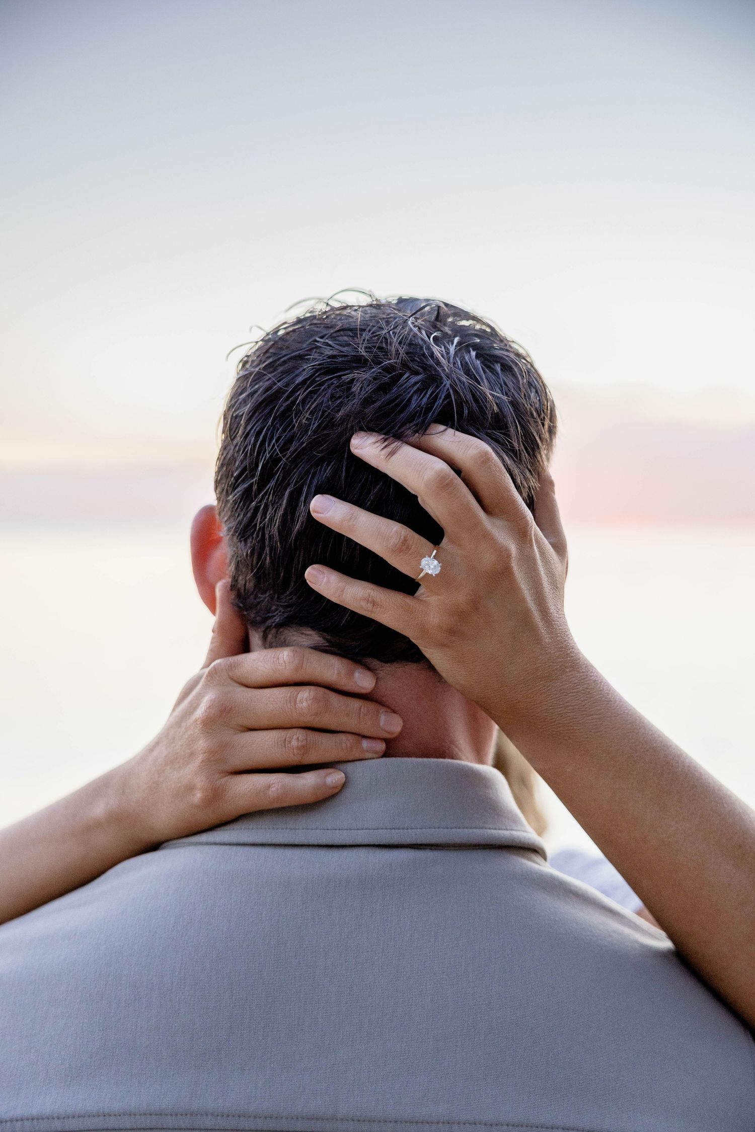 A woman with an engagement ring tenderly holds a mans head from behind at sunset.
