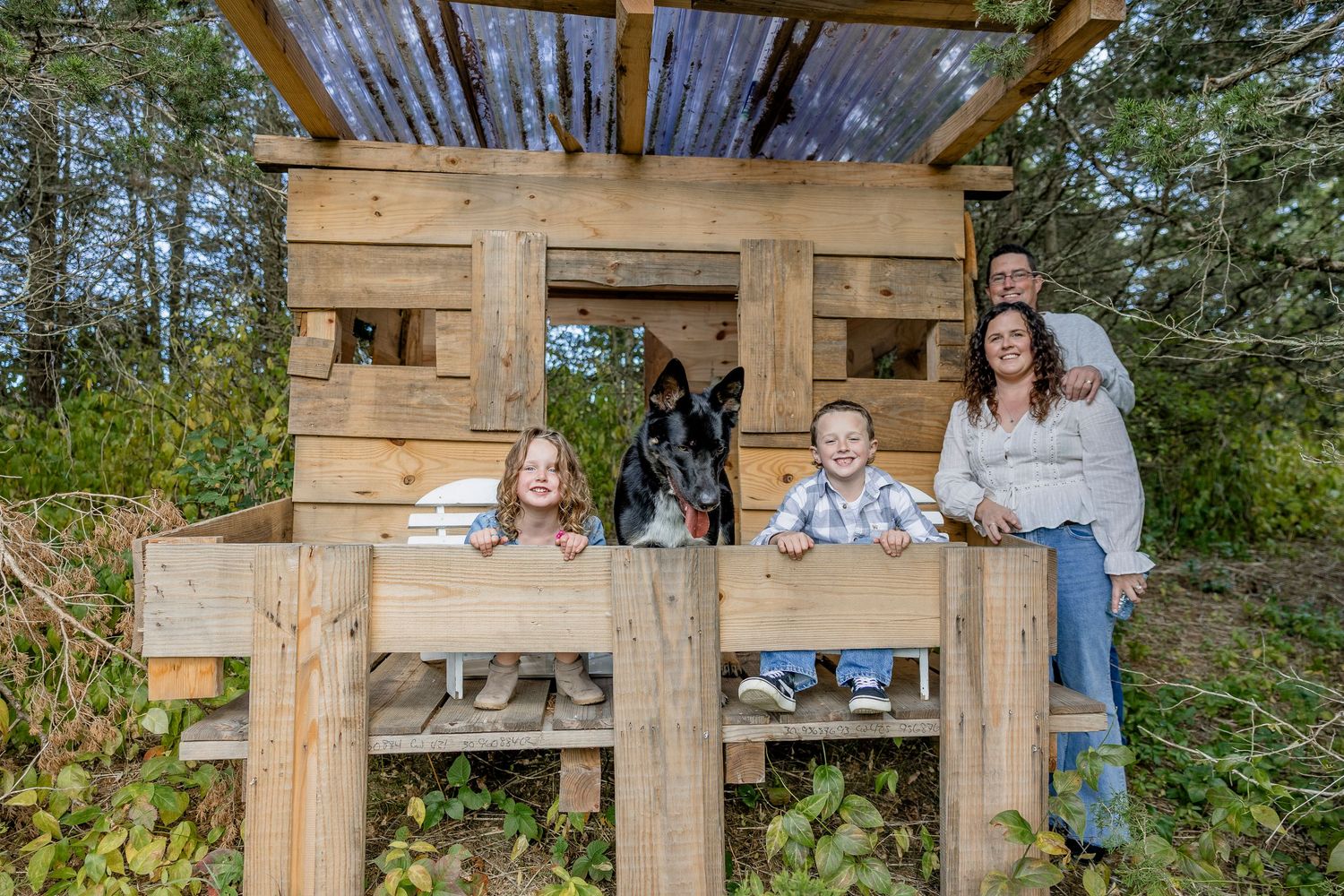 Family with dog posing inside a rustic wooden pallet playhouse structure outdoors surrounded by trees.
