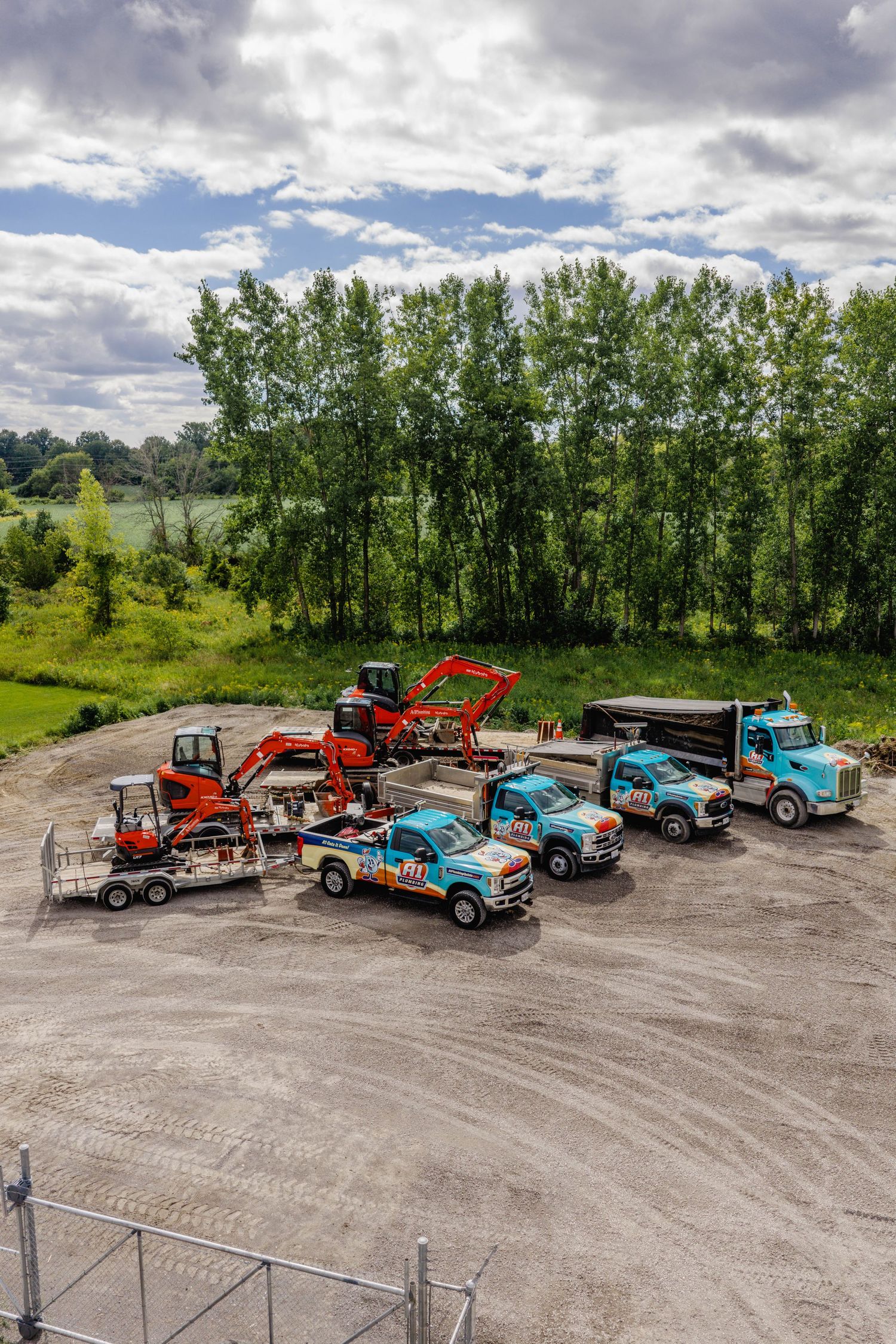 Aerial view of blue construction trucks and orange excavator parked on gravel lot surrounded by lush green trees.