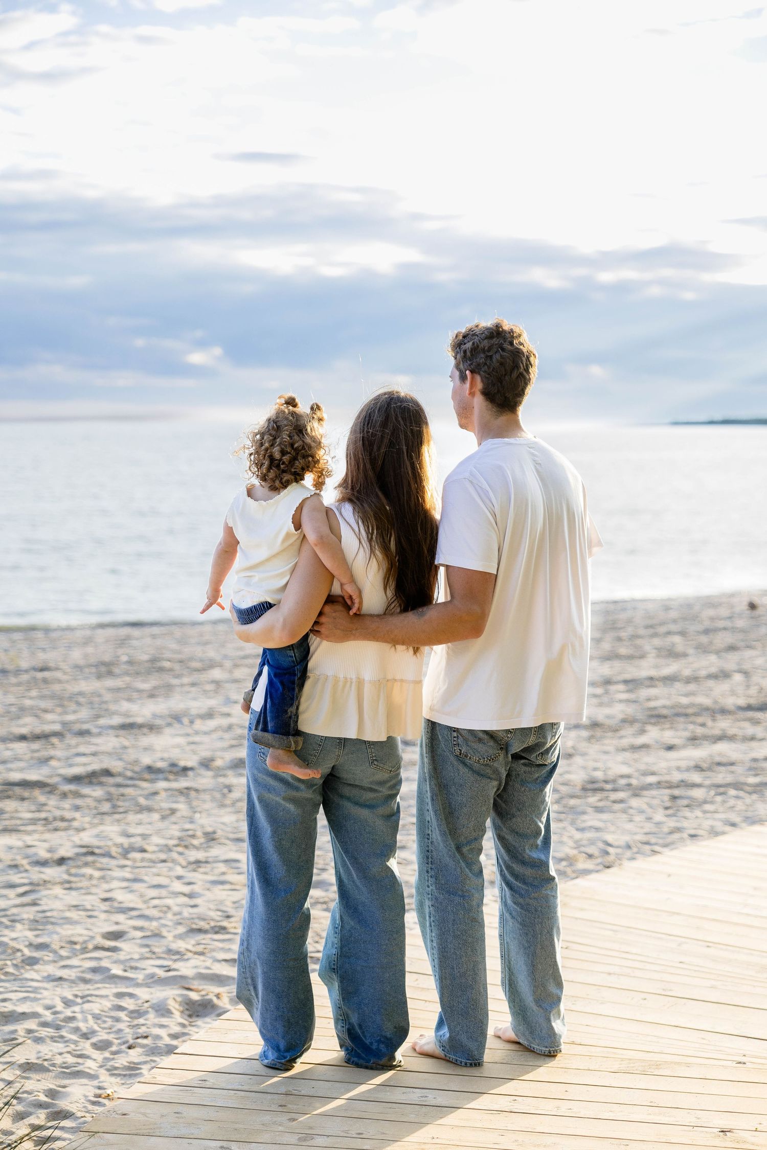 Family of three viewed from behind on beach boardwalk, parents embracing child while gazing at ocean.