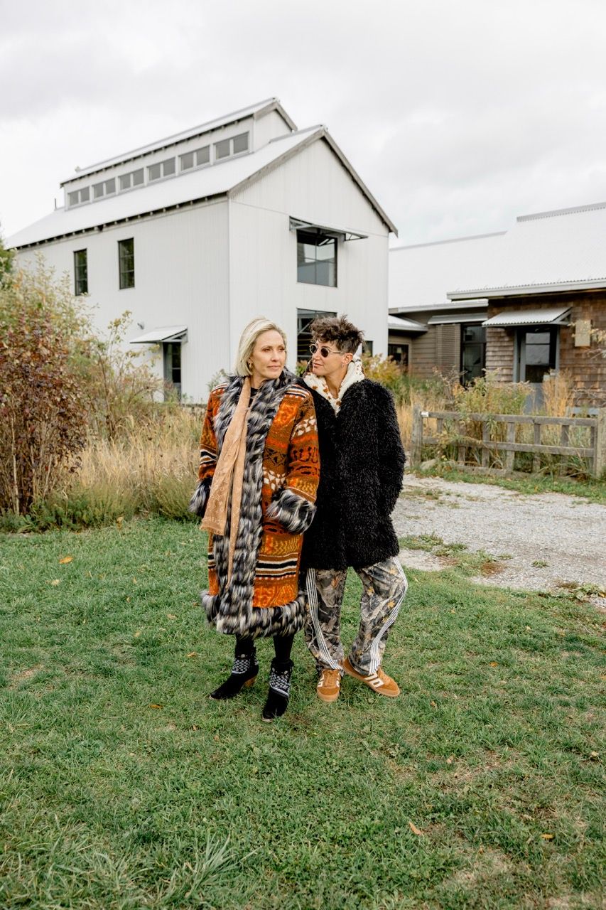 Two fashionably dressed individuals stand on a grassy lawn in front of a modern white farmhouse.